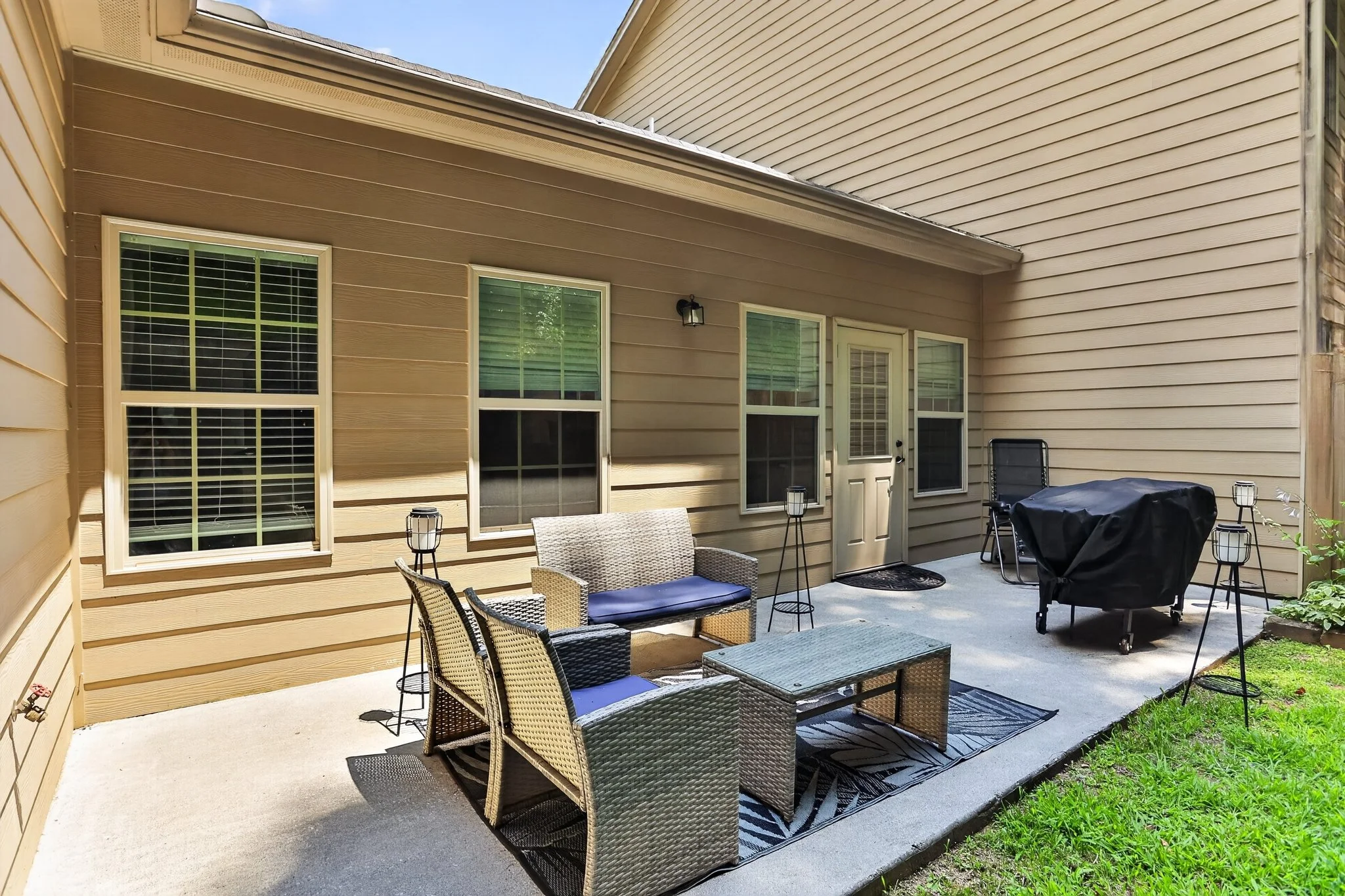 Backyard patio with outdoor furniture including a wicker seating set with cushions, a table, and a grill covered with a black cover. There are two small lanterns on the patio floor. The patio is surrounded by beige house siding, with three windows and a door visible. There is a small patch of grass and greenery at the edge of the patio.