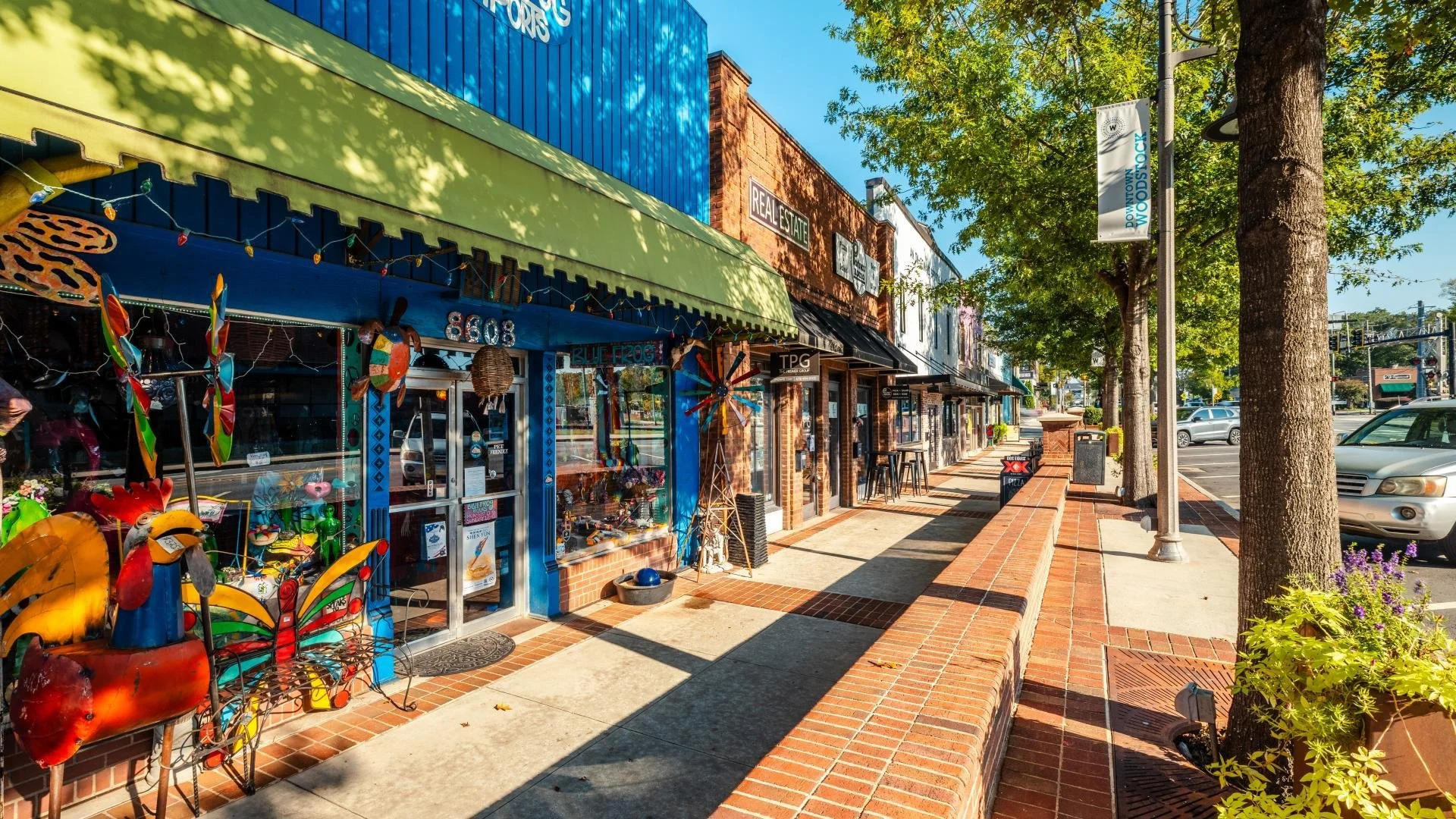 A colorful storefront on a sidewalk in a small town or shopping district with trees, parked cars, and a blue sky.