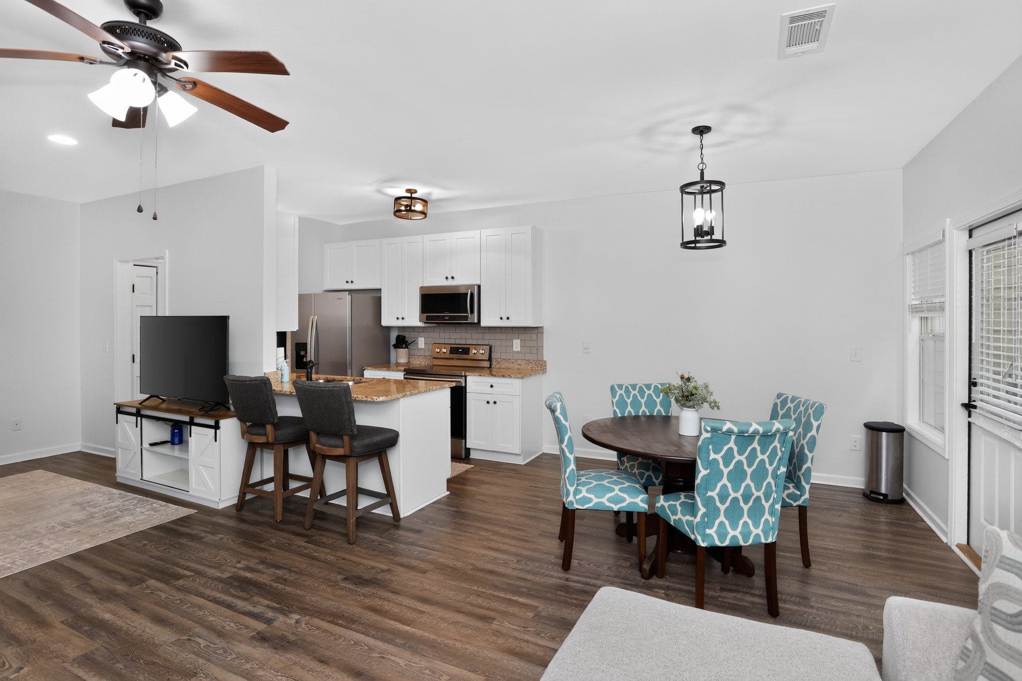 Open-concept kitchen and dining area with white cabinets, stainless steel appliances, and a round wooden table with four blue patterned chairs. A ceiling fan, pendant light, and small ceiling lights illuminate the space. The floor is dark wood, and there is a window with blinds on the right.