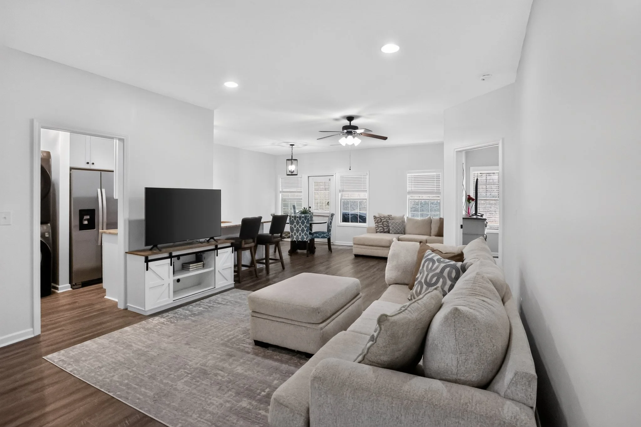 Living room with a beige sofa, patterned pillows, a TV on a stand, a dining table with chairs, and multiple windows with blinds, illuminated by ceiling lights and a ceiling fan.