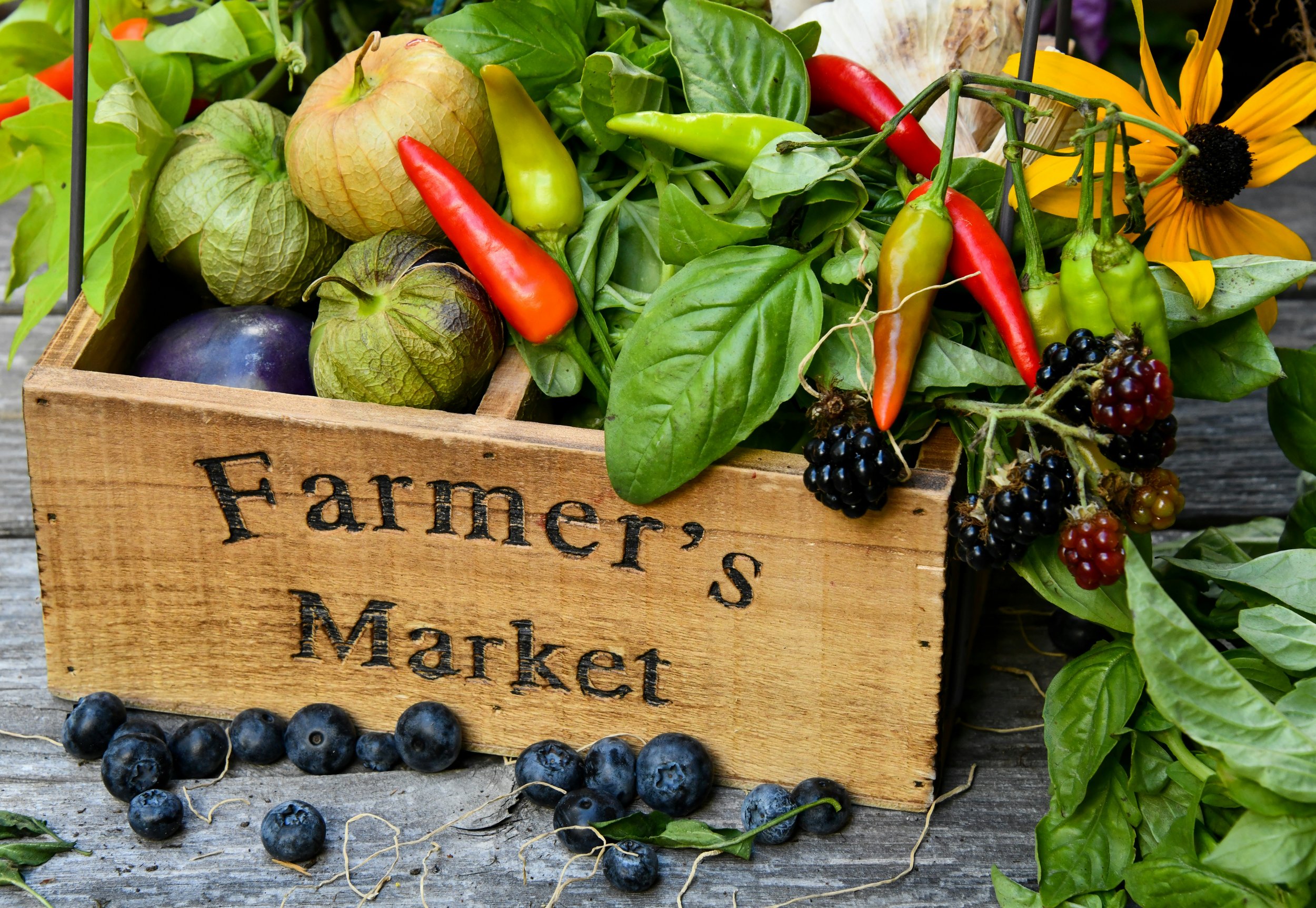 A wooden crate filled with fresh fruits and greens, including apples, peppers, eggplant, berries, and leafy vegetables, with some spilling onto the rustic wooden surface below.