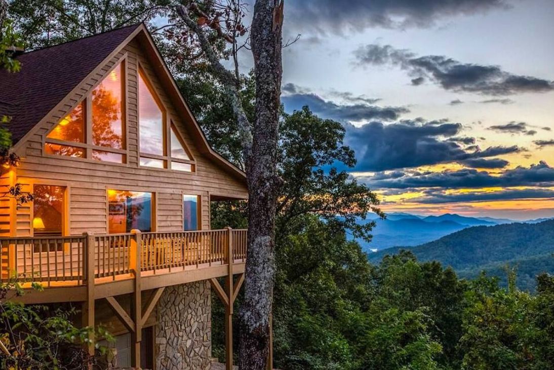 A wooden house on a mountain with large windows, surrounded by trees, overlooking a scenic landscape of mountains during sunset.