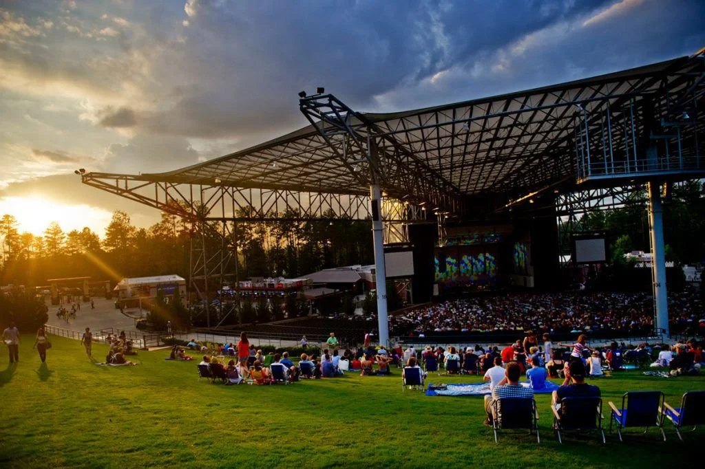 An outdoor concert at sunset with people sitting on grass and in chairs, a large stage with a canopy, and a forested background.
