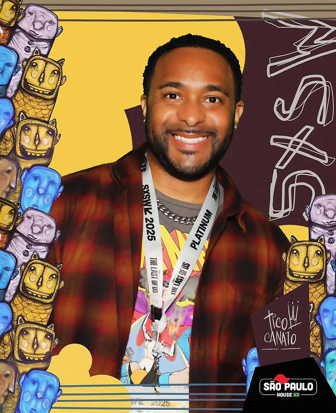 A man smiling in front of a colorful backdrop with cartoonish characters; he is wearing a lanyard with event information and a casual outfit.