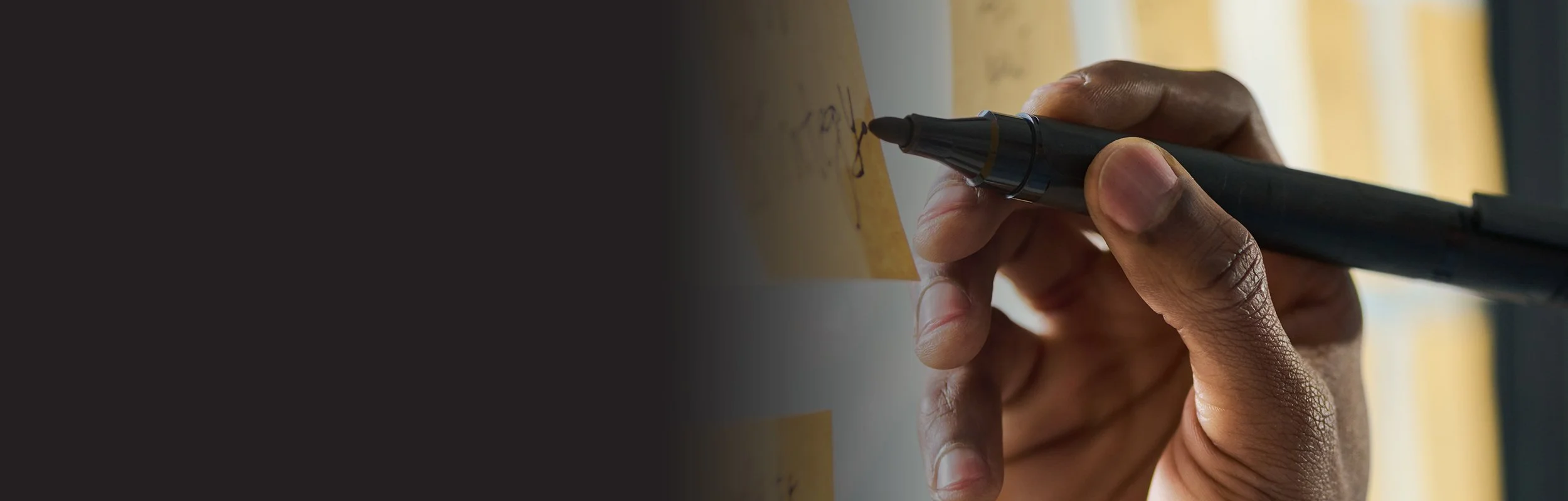 A person holding a black pen and writing on a sticky note.