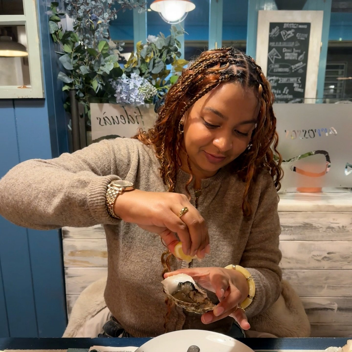 A woman with braided hair and gold jewelry opening an oyster in a restaurant with blue decor, plants, and a chalkboard menu in the background.