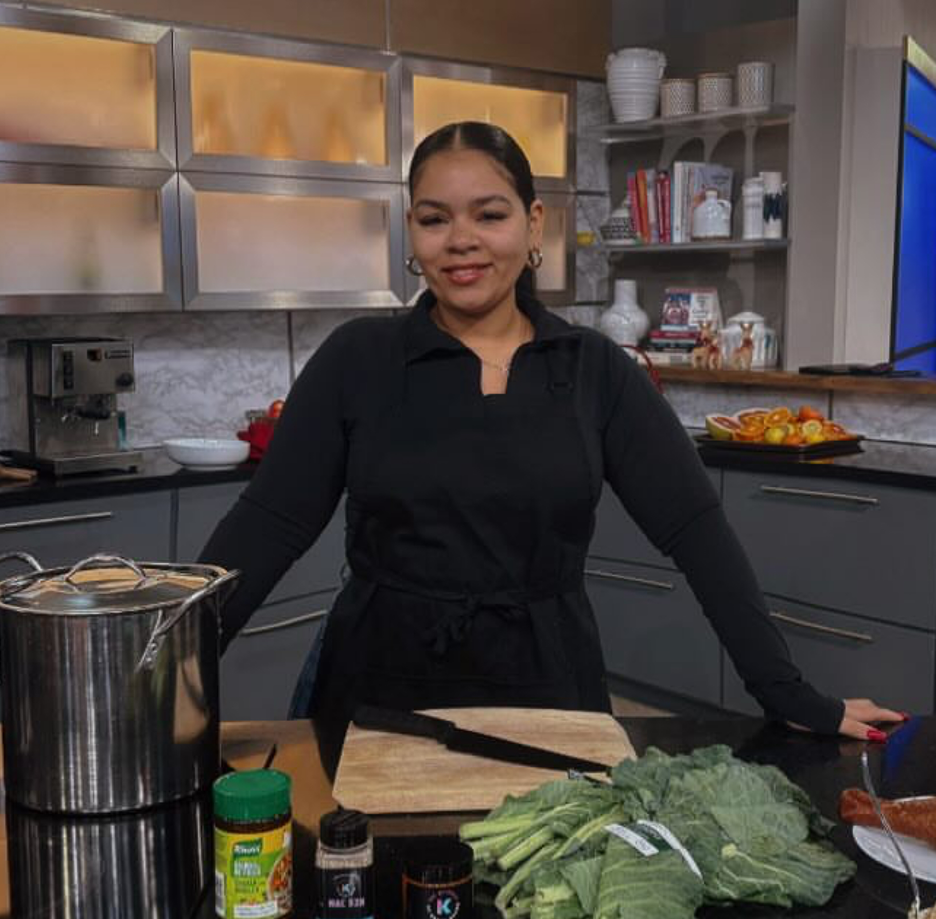 A woman wearing a black shirt inside a kitchen, surrounded by various food ingredients including vegetables, canned goods, and spices.