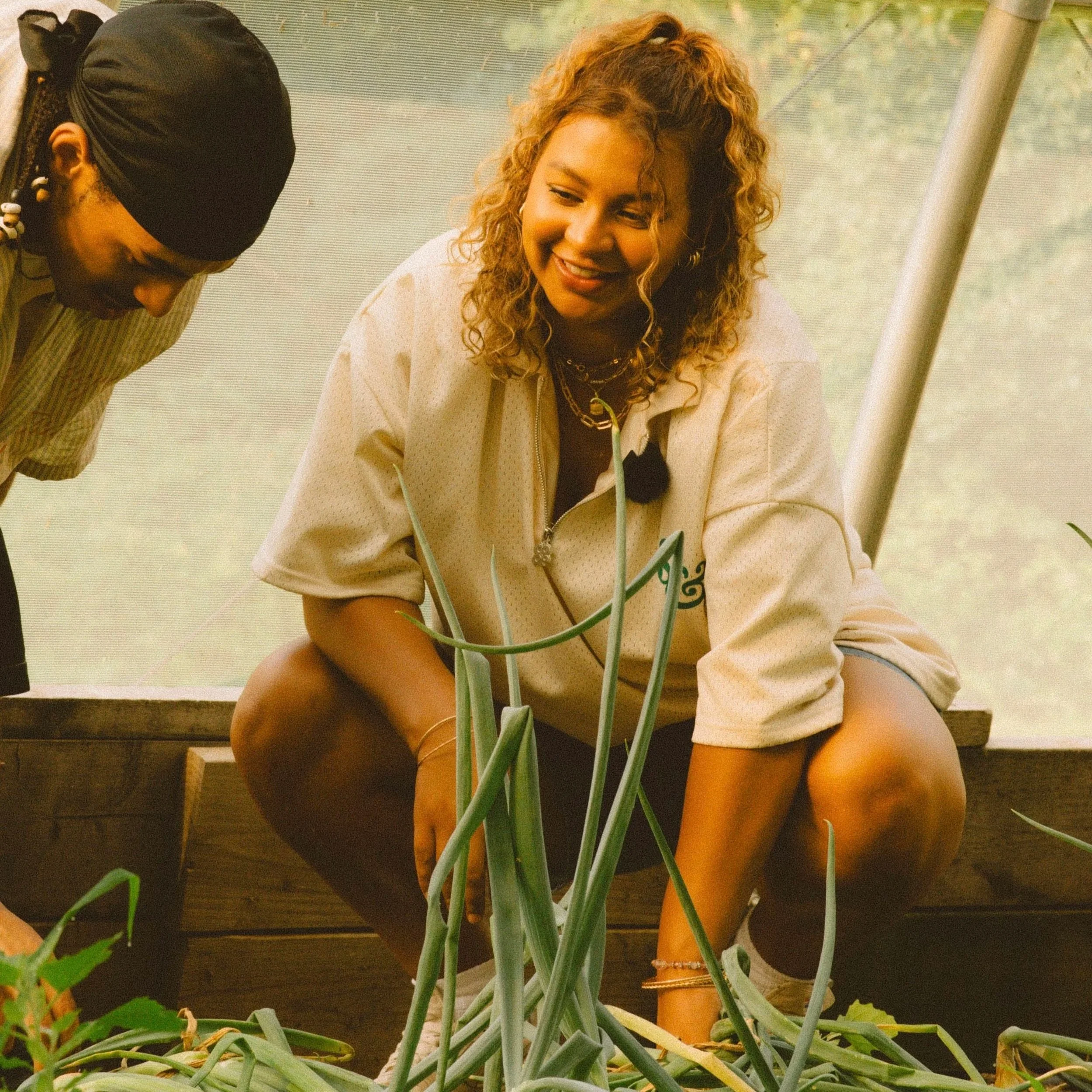 A woman with braided hair and gold jewelry opening an oyster in a restaurant with blue decor, plants, and a chalkboard menu in the background.