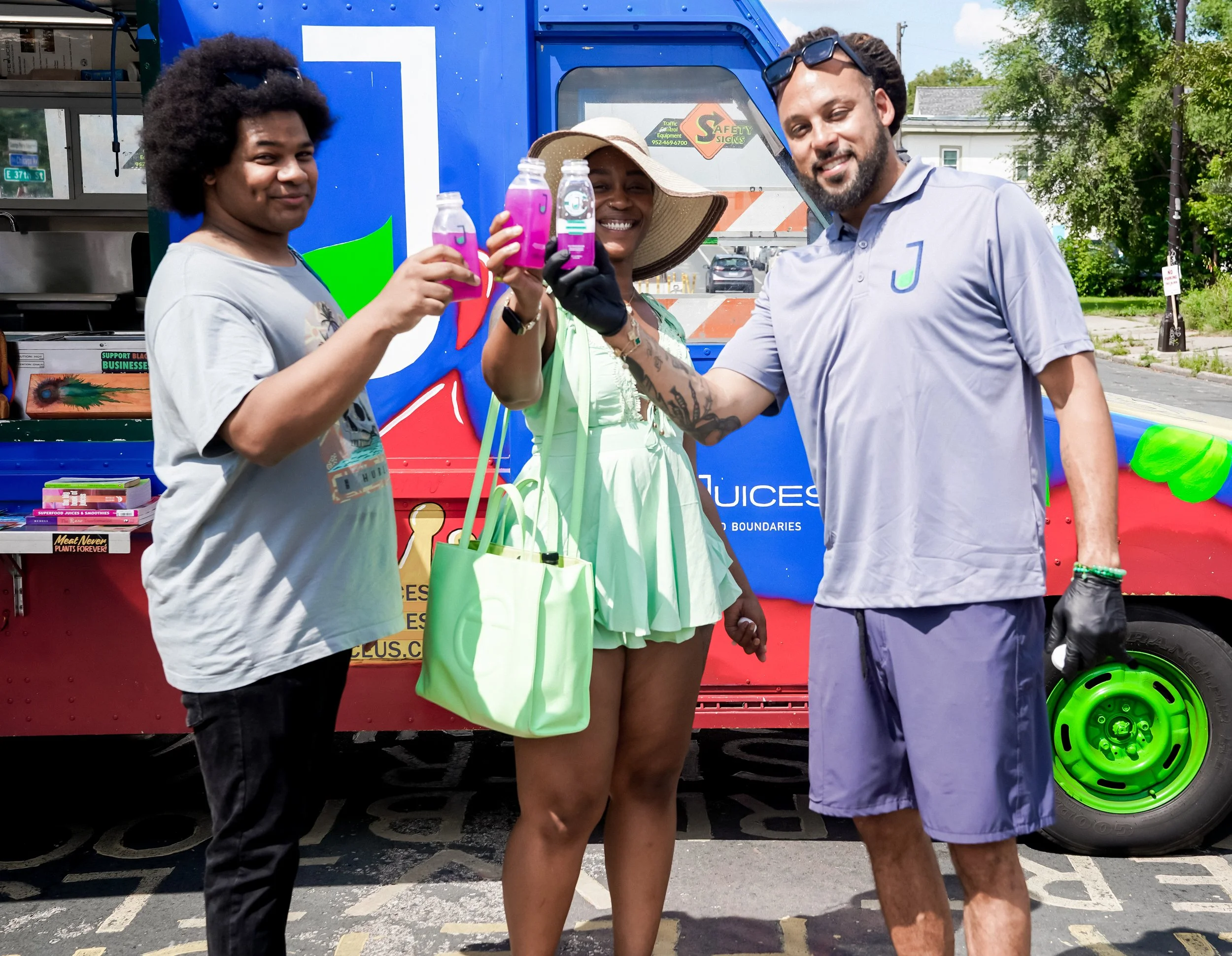 Three people standing in front of a blue juice truck, holding bottles of pink juice, smiling.