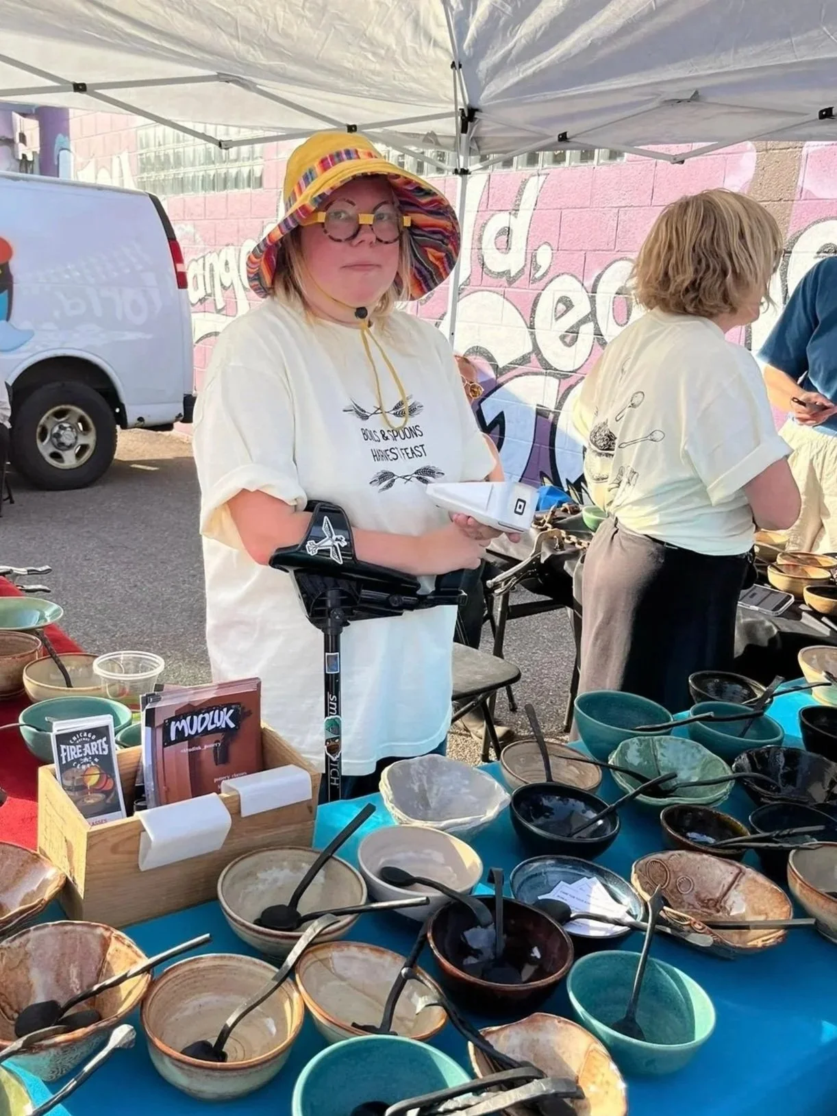 A woman wearing a colorful sun hat, glasses, and a white t-shirt standing behind a table with various bowls and utensils at an outdoor market. Another woman is visible in the background, and a van is parked nearby.