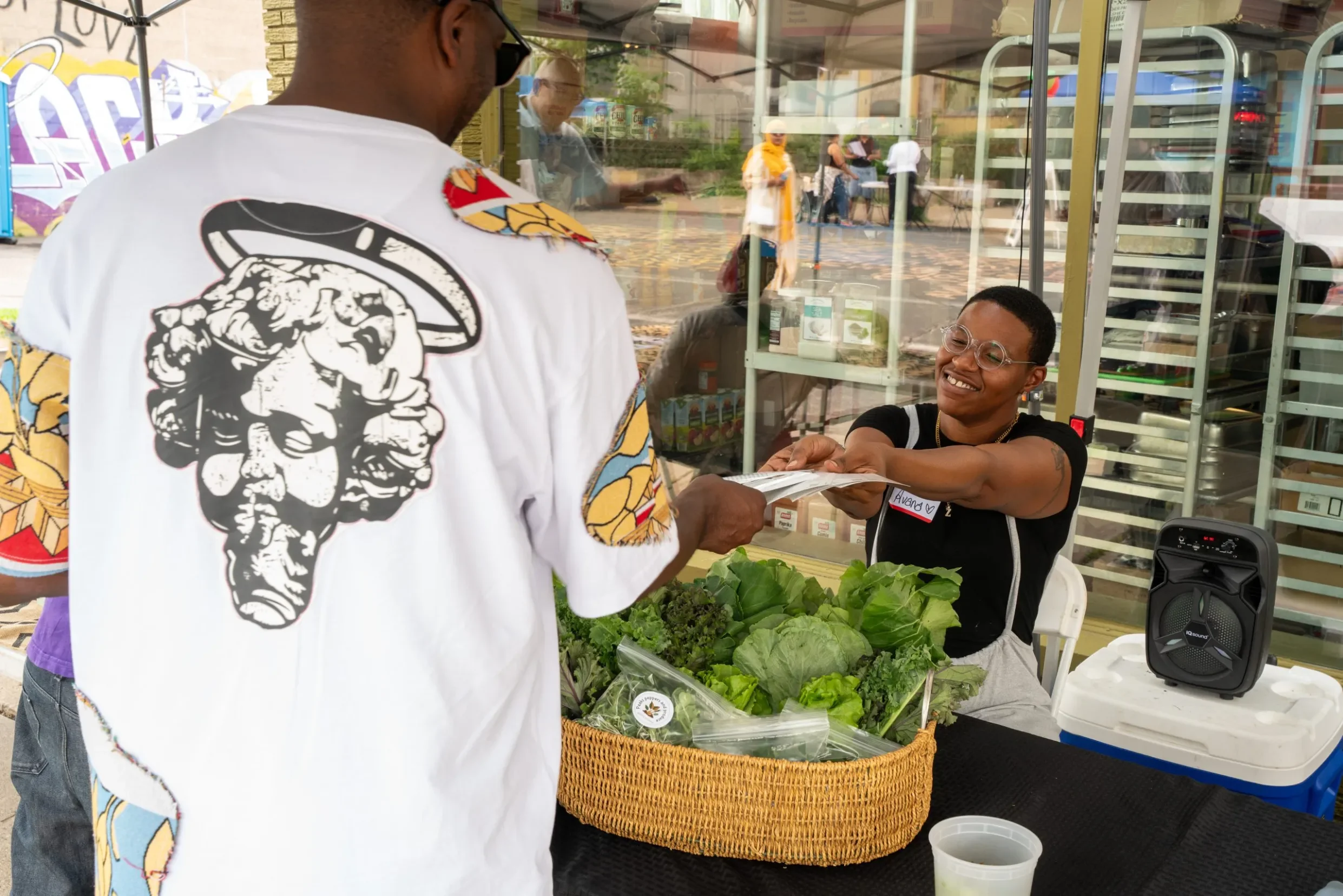 A man in a white graphic t-shirt is at a produce stand, handing money to a smiling woman with glasses behind the counter. The stand has leafy greens and other vegetables. Reflections of people and outdoor scene are visible on the glass display case.