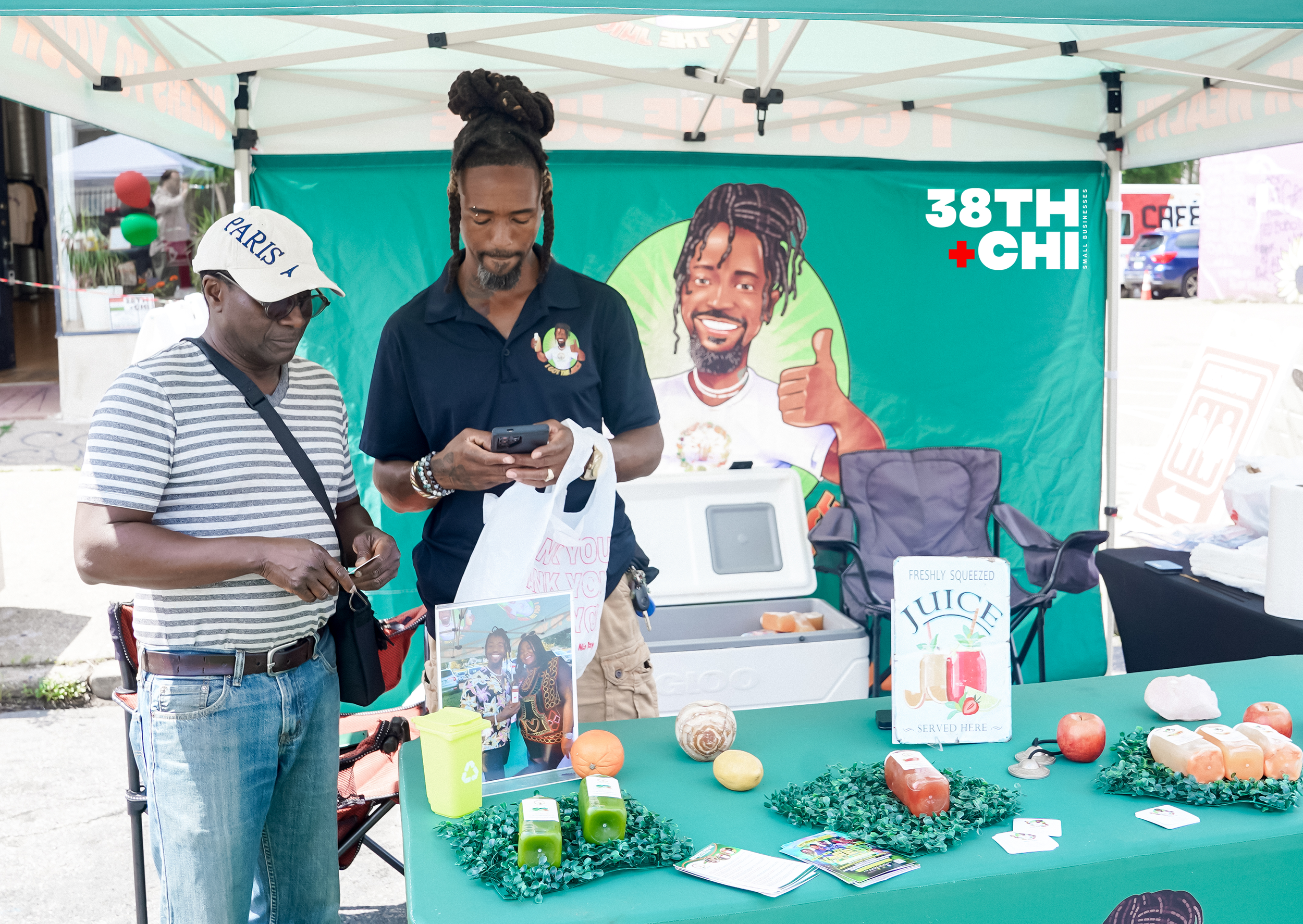 Two men are at a booth selling produce at an outdoor market under a white canopy tent. The table displays apples, pumpkins, and bottled juice, with a sign reading "Freshly Squeezed Juice." One man is wearing a striped shirt, a white cap, and glasses and is looking at his phone. The other man, with dreadlocks tied up, is wearing a dark polo shirt and is also looking at his phone. A colorful banner with a smiling cartoon character and the text "38TH + CHI" is in the background.