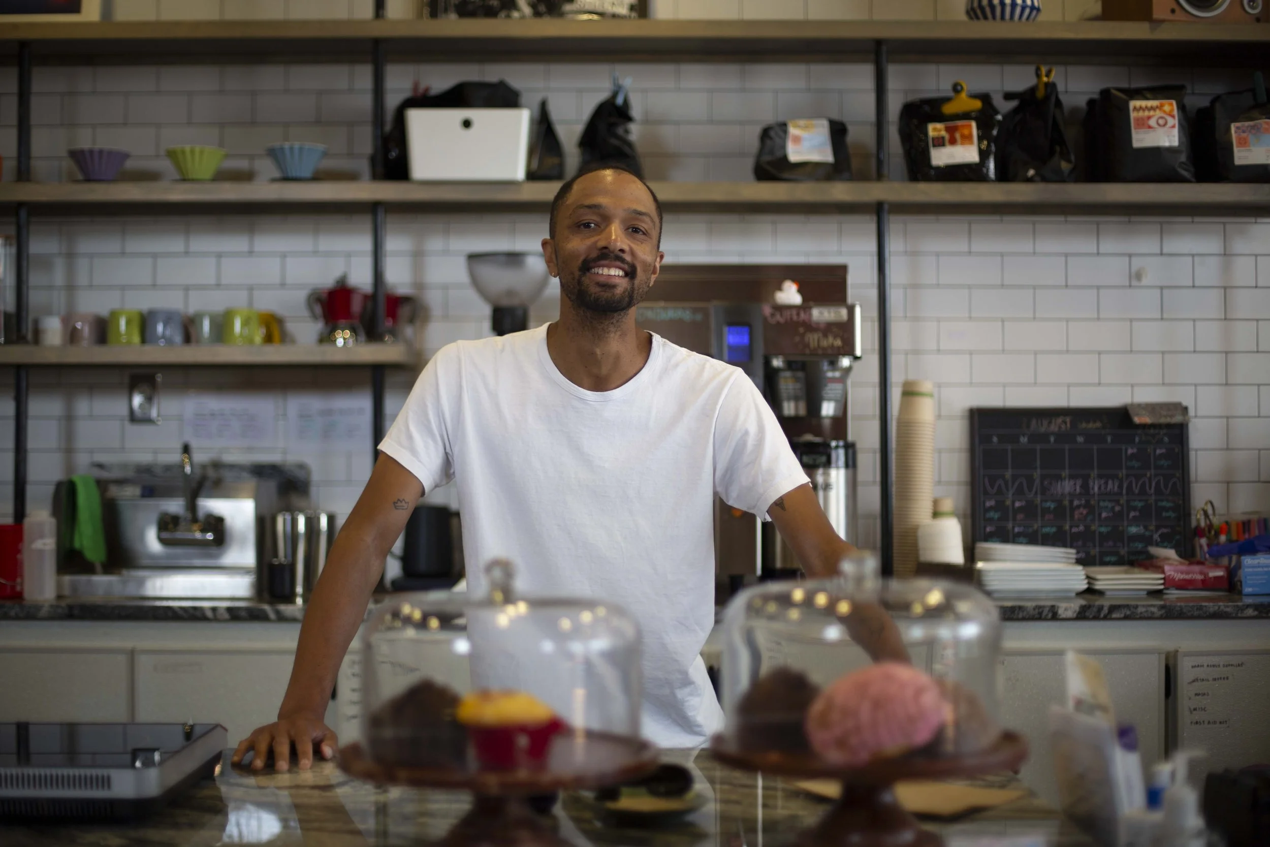 A man with a beard and short hair, wearing a white t-shirt, standing behind the counter in a cafe or bakery. Shelves with colorful cups and bags of coffee beans are visible in the background. There are baked goods and cups on the counter in the foreground.