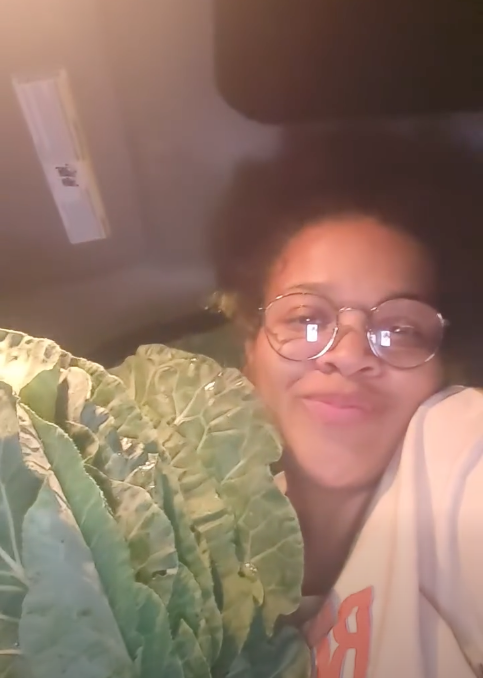 A woman with glasses smiling and lying down next to a large leafy green plant in a car.