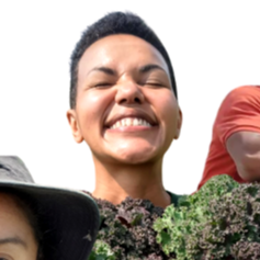 Close-up of a smiling woman with short hair outdoors, surrounded by greenery and other people.