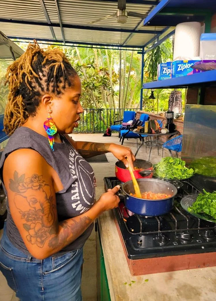 Woman with colorful earrings and tattoos cooking on a stove in an outdoor kitchen, with leafy plants in the background.