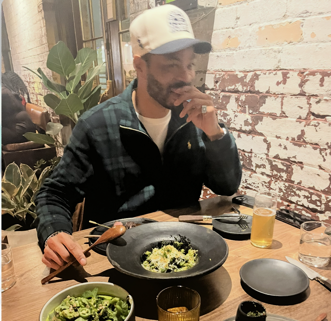 A man sitting at a table in a restaurant, wearing a cap, checking his phone, with plates of food and drinks on the table, including a bowl of salad, a glass of beer, and a plate of pasta.