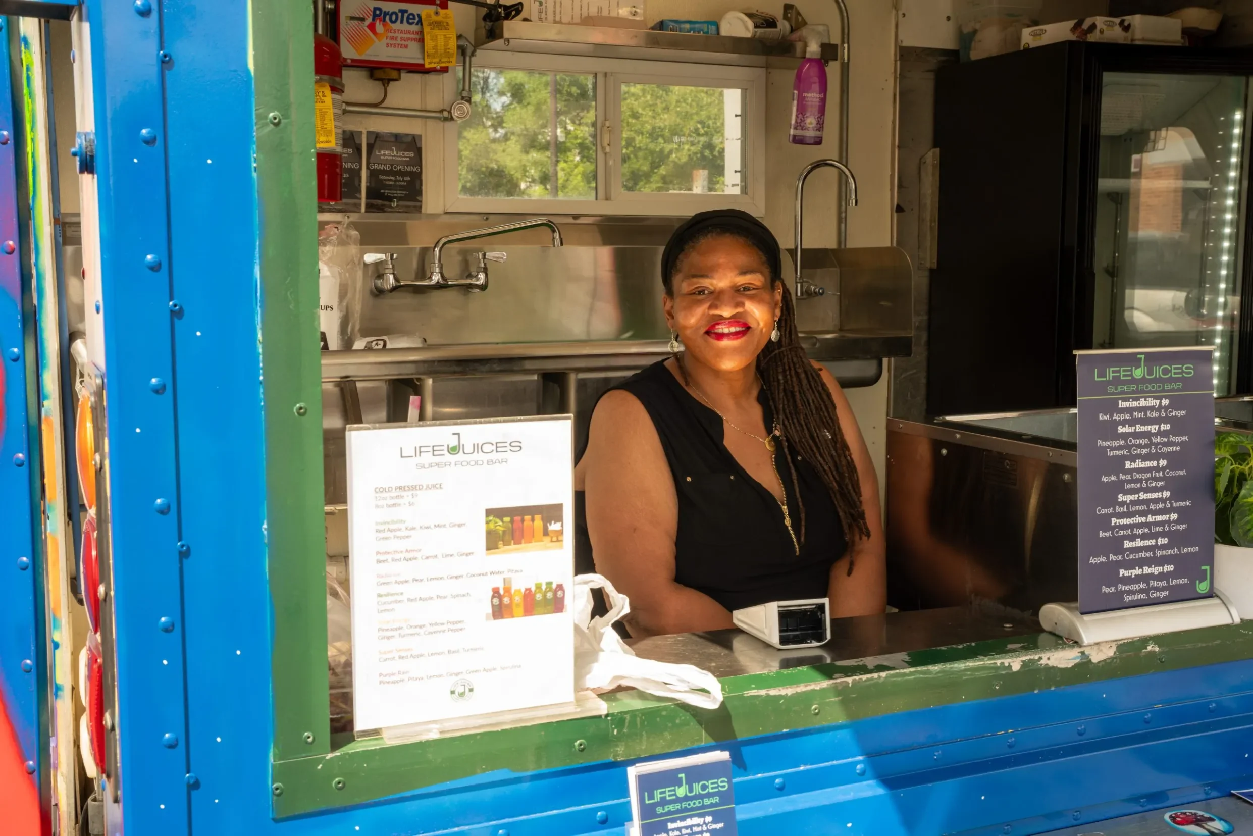 A woman smiling behind a food stand at a juice bar called 'LIFEJUICES Super Food Bar' with a window, and various juice options listed on a sign in front of her.