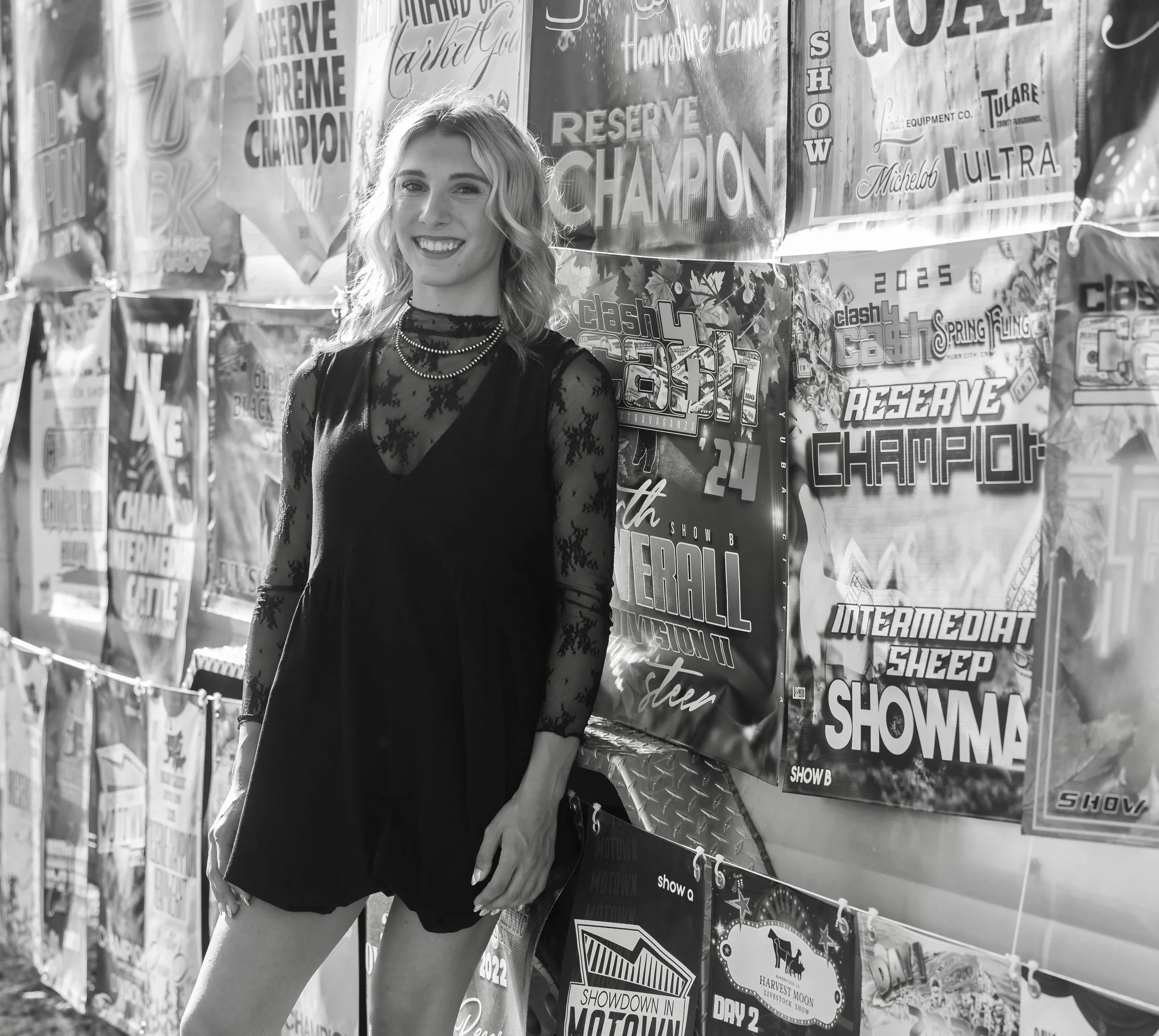 A young woman with blonde, wavy hair smiling in front of a wall covered with various event posters and signs.