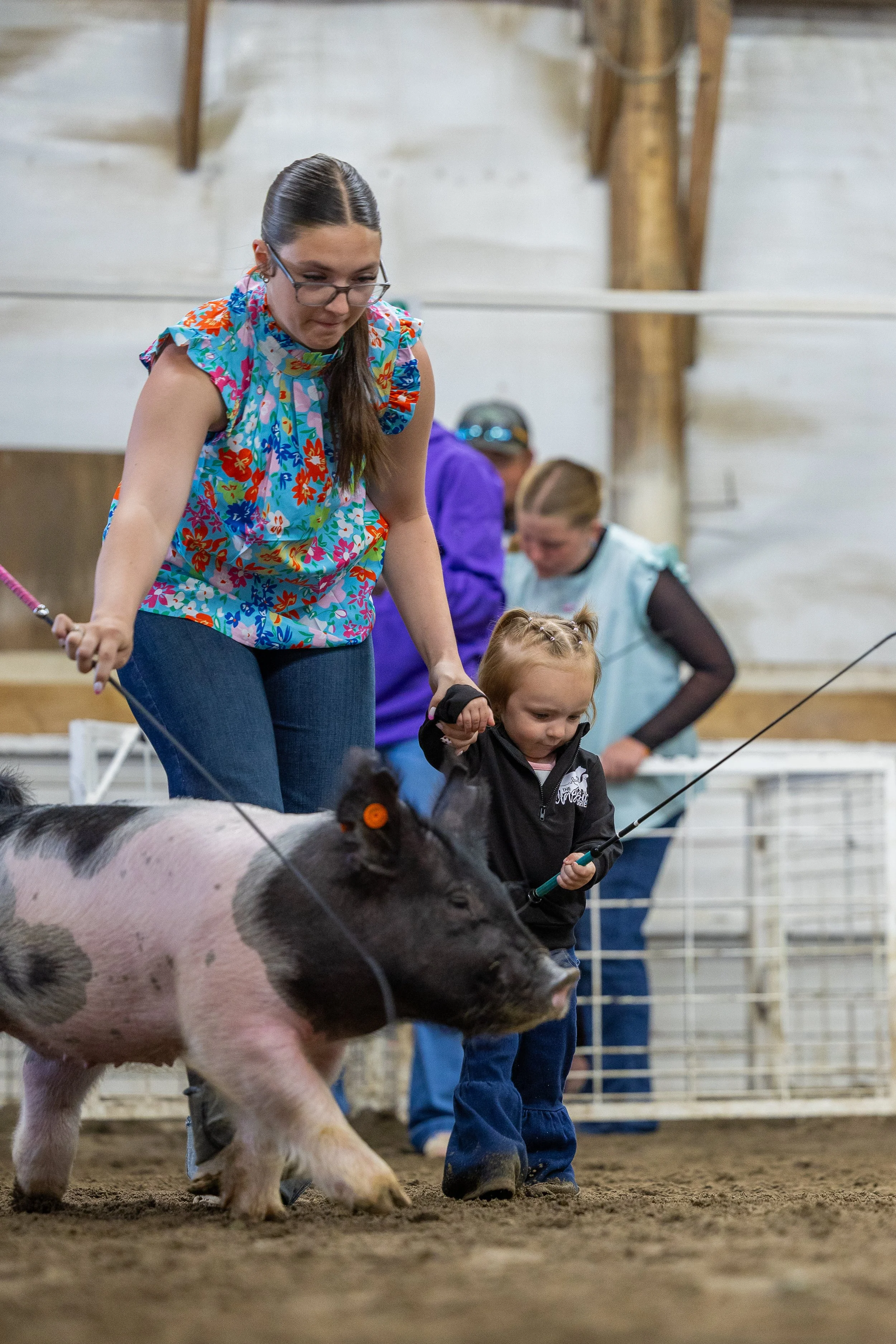 A young girl and an adult woman are at a pig show, holding a pig by its leash inside a barn or indoor arena. The girl is wearing a black jacket and the woman is wearing glasses and a colorful, floral sleeveless shirt.
