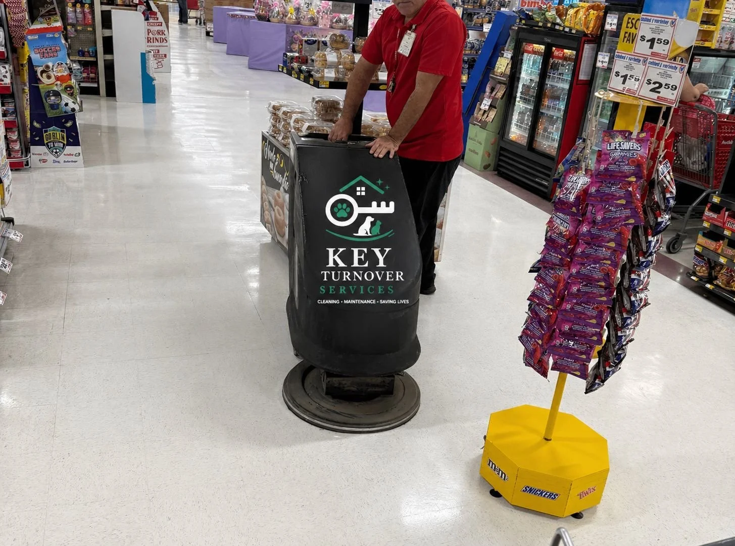 A person in a red shirt is standing behind a black cart that has the logo and name of 'Key Turnover Services' on it, in a grocery store aisle. The store has various products on shelves, including snacks and baked goods, and a display rack of purple snack bags on the right. There is a bright yellow stand with snack packaging on the right side of the person.