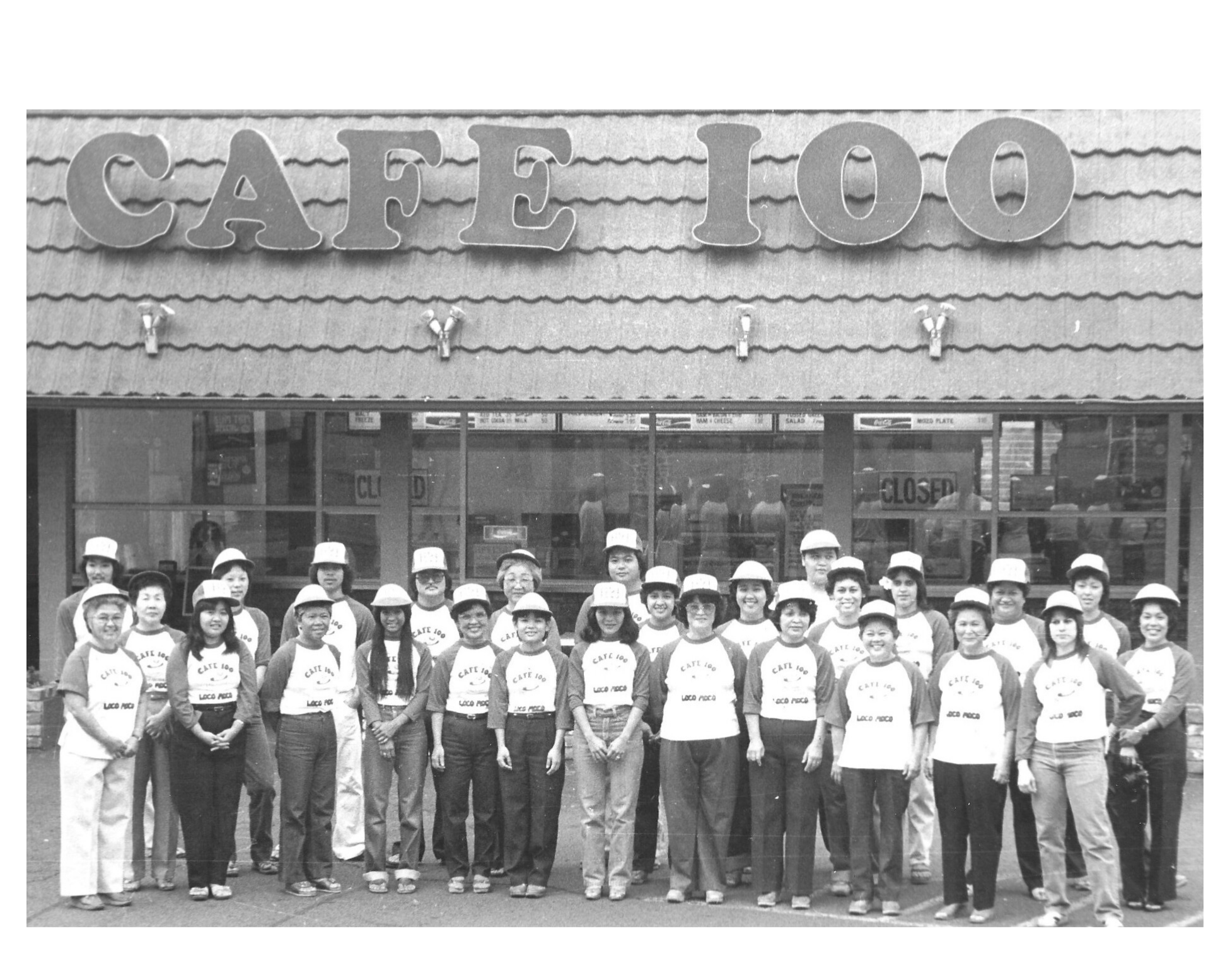 A group of people posing in front of a cafeteria with a sign that says 'Cafe 100'. Some are wearing matching T-shirts and hats, indicating they are staff or part of a team.