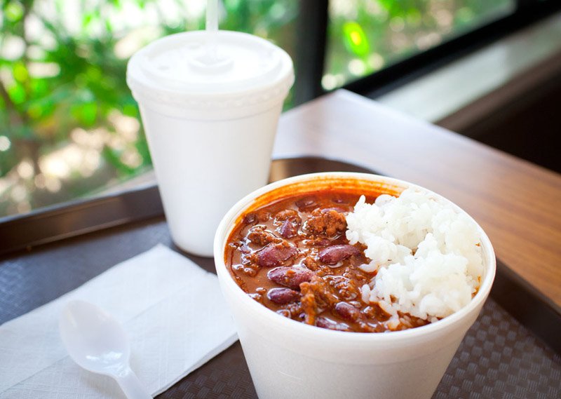 A cup of chili with beans and rice, a white disposable cup with a lid, and a plastic spoon on a napkin on a tray at a restaurant.