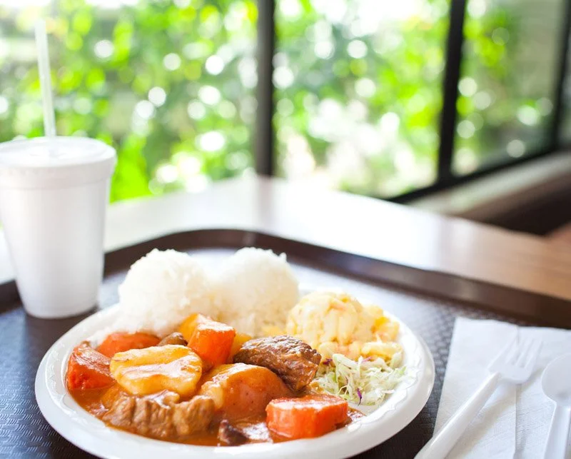 Plate of stew with carrots and beef, mashed potatoes, potato salad, and coleslaw on a table near a window