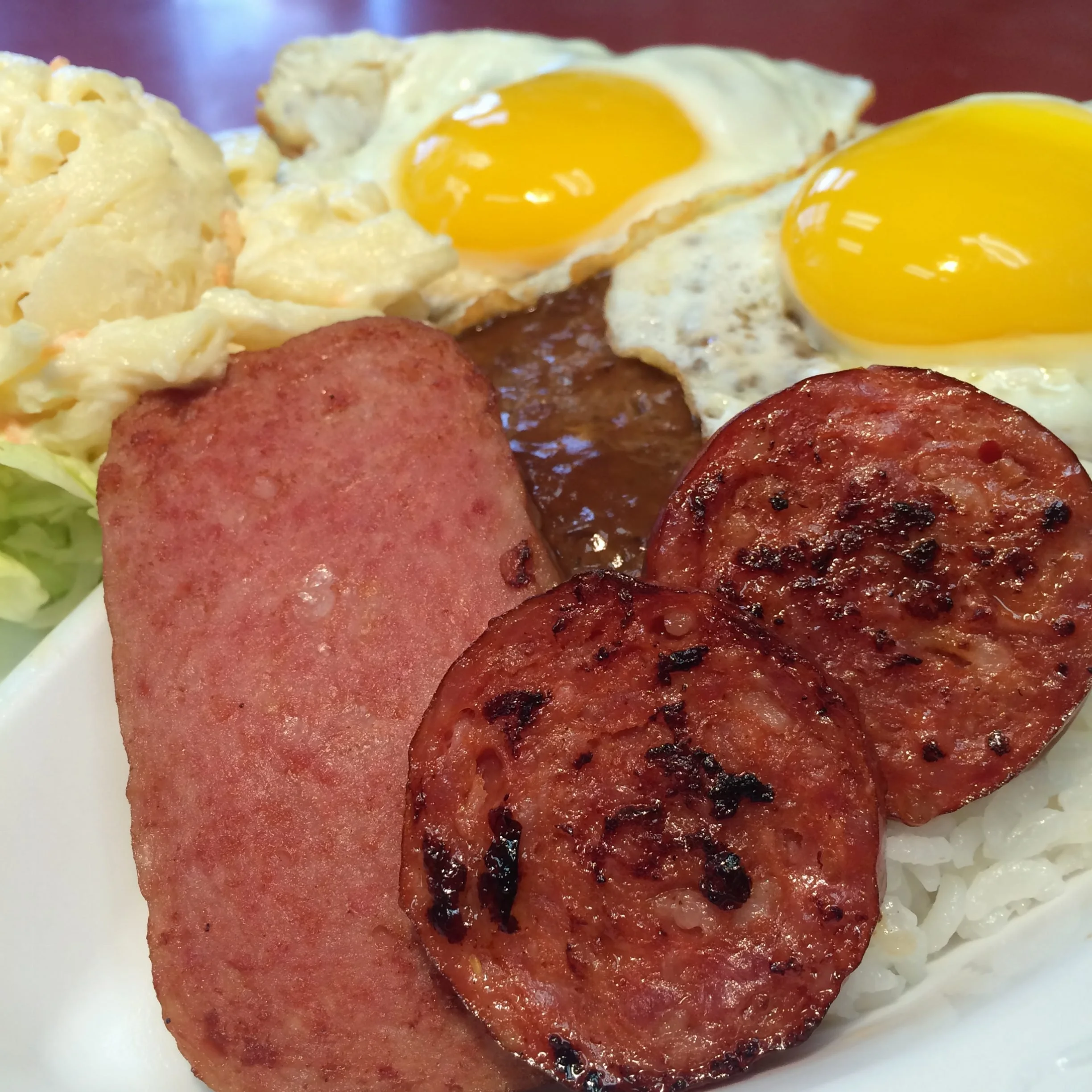 Breakfast plate with scrambled eggs, two sunny-side-up eggs, grilled ham and sausage, rice, and potato salad.