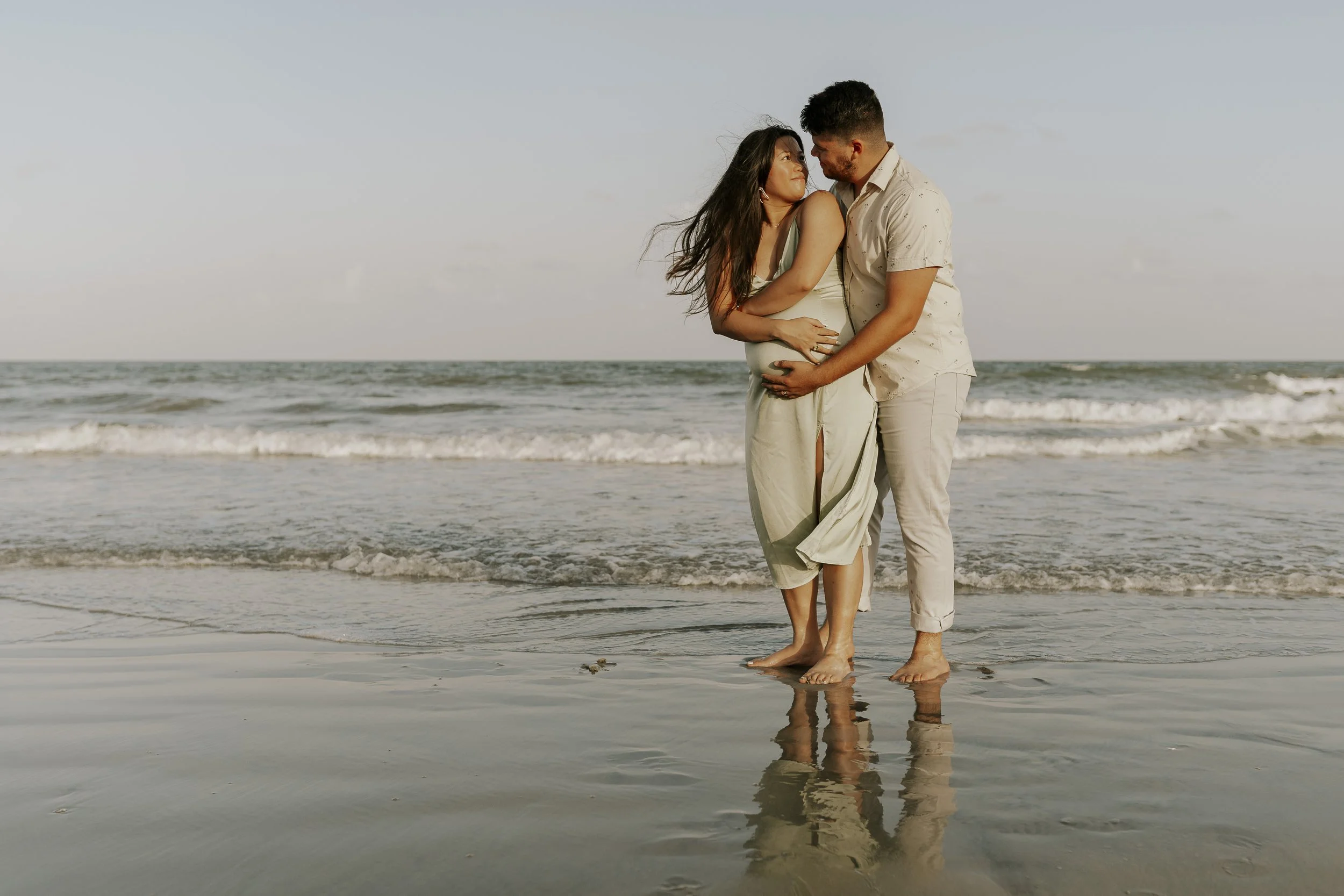 A couple standing on the beach, touching noses, with the ocean waves behind them, during sunset or late afternoon.