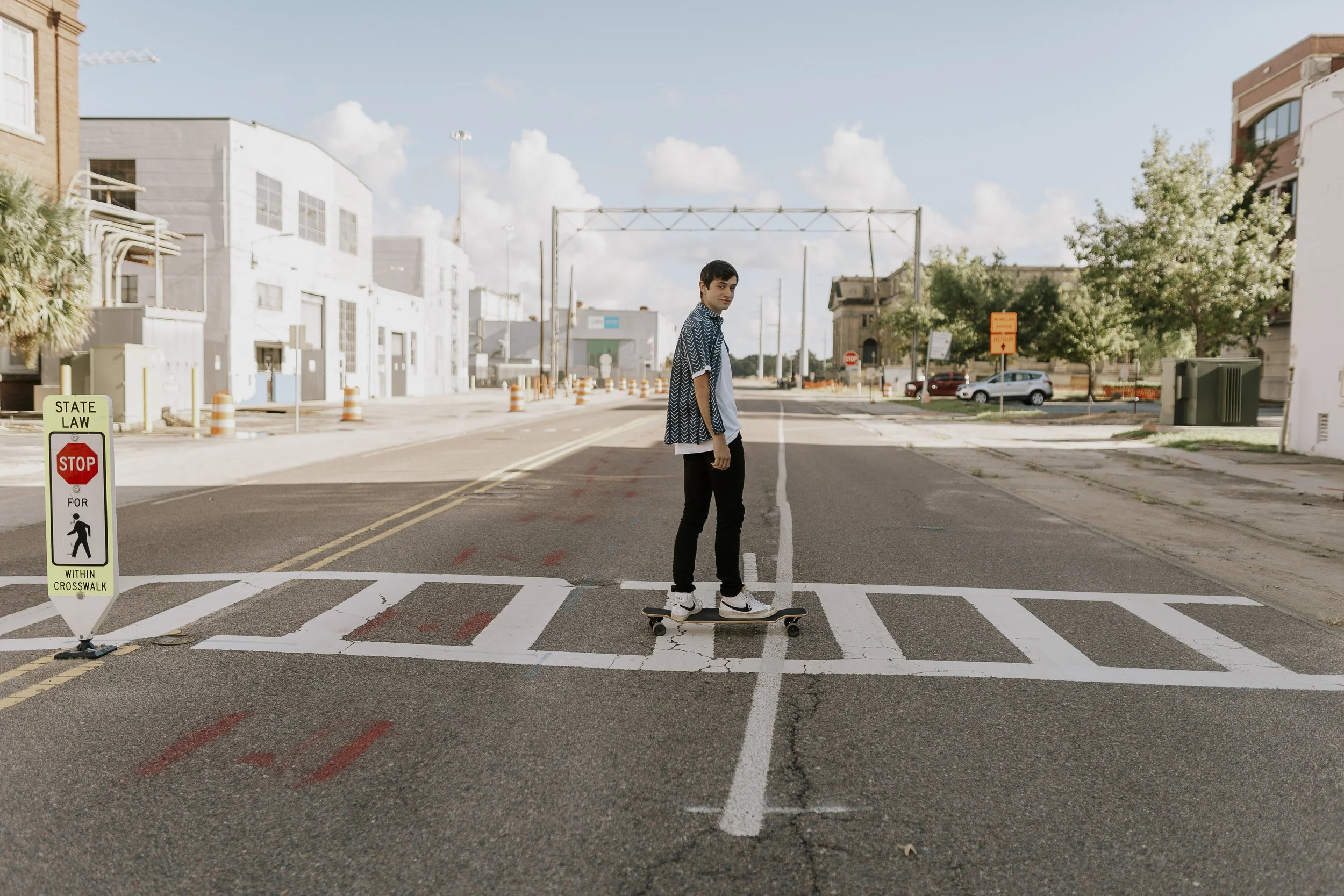 A young man skateboarding across a marked crosswalk in an urban area during daytime. There are white buildings, cars parked, trees, and orange construction cones along the street. A sign in the foreground indicates the crosswalk.