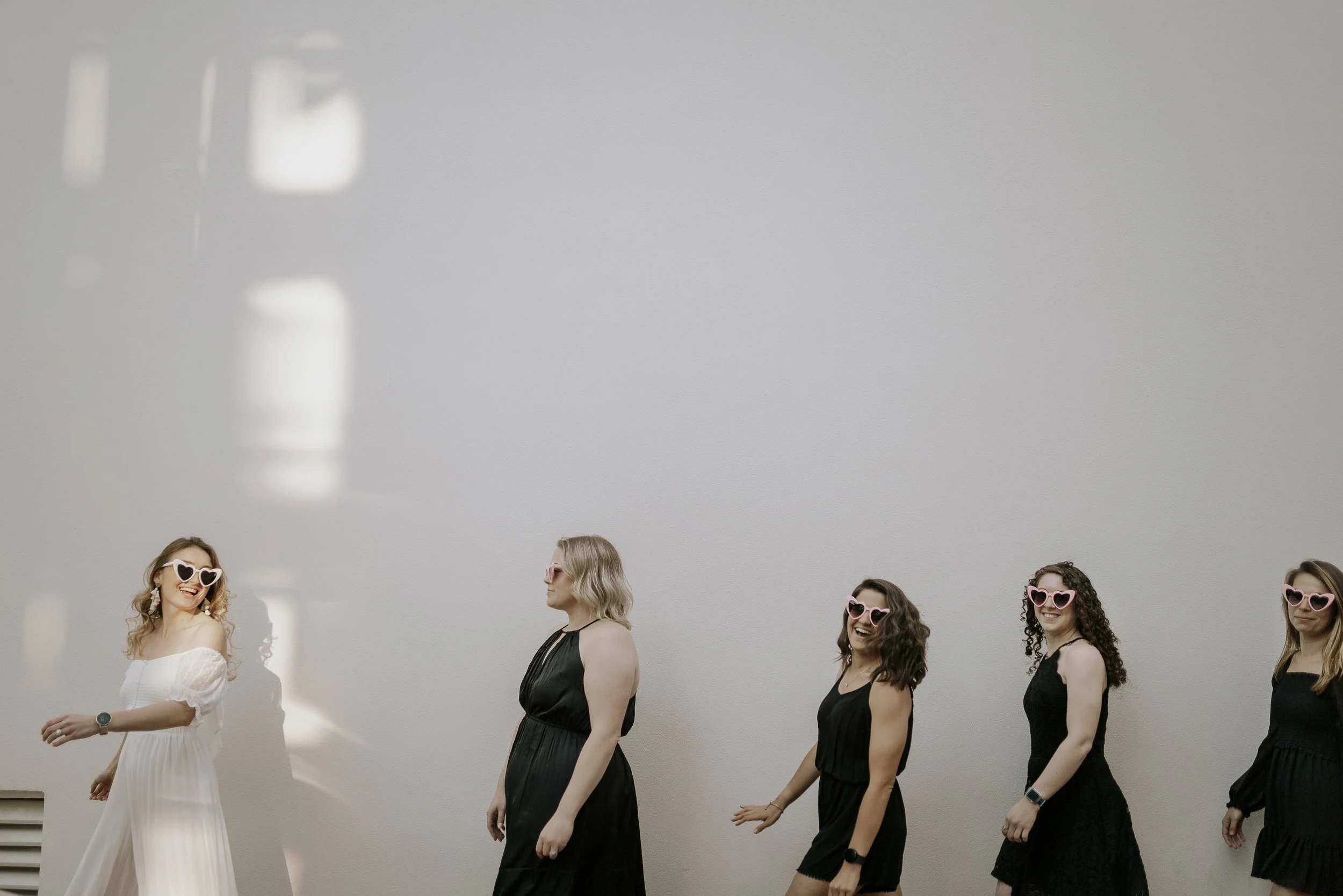 A group of women walking in a line, wearing black dresses and heart-shaped sunglasses, with one woman in a white dress and heart-shaped sunglasses smiling, against a plain gray wall.