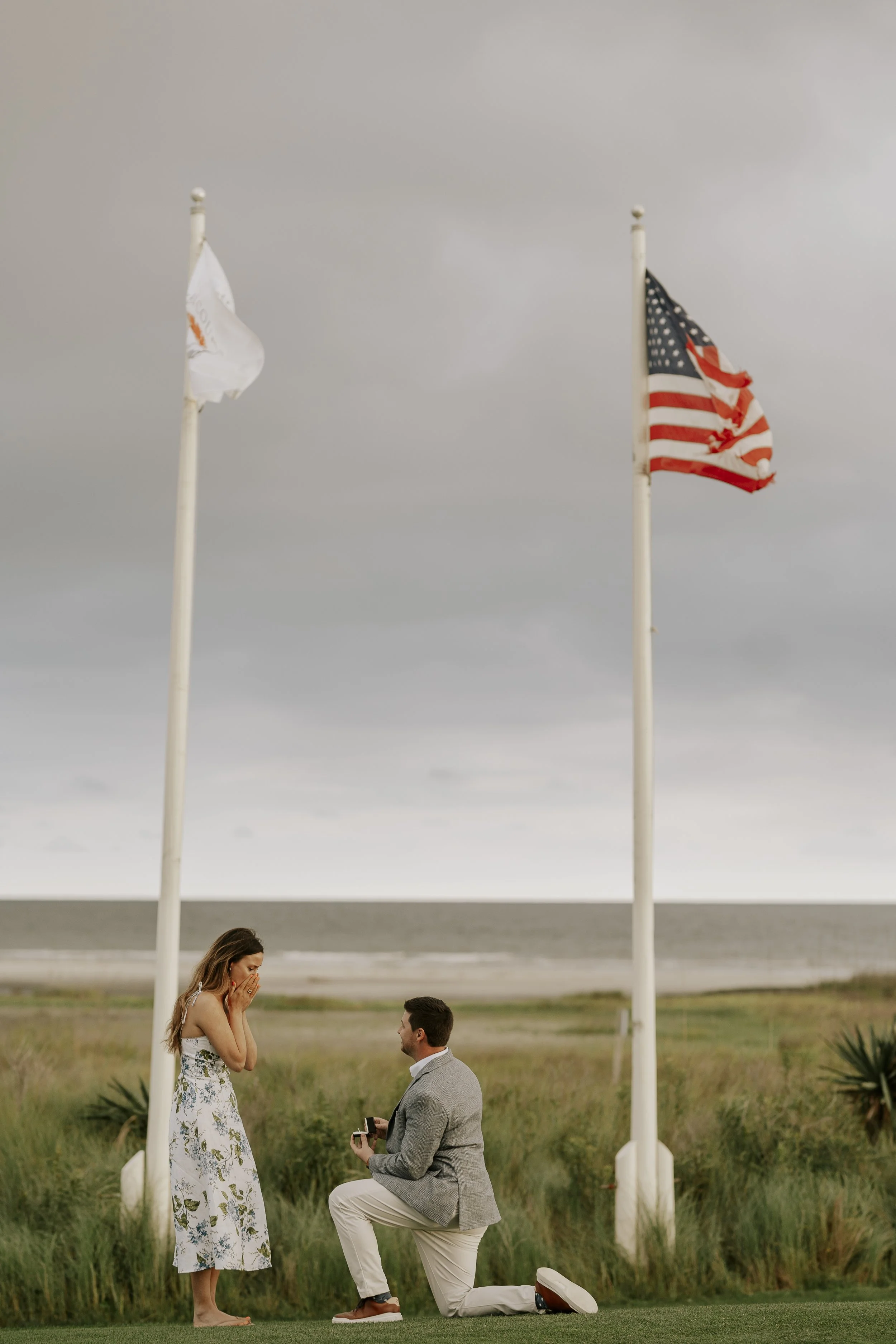 A man is kneeling on one knee proposing marriage to a woman in a field, with two American flags flying in the background.