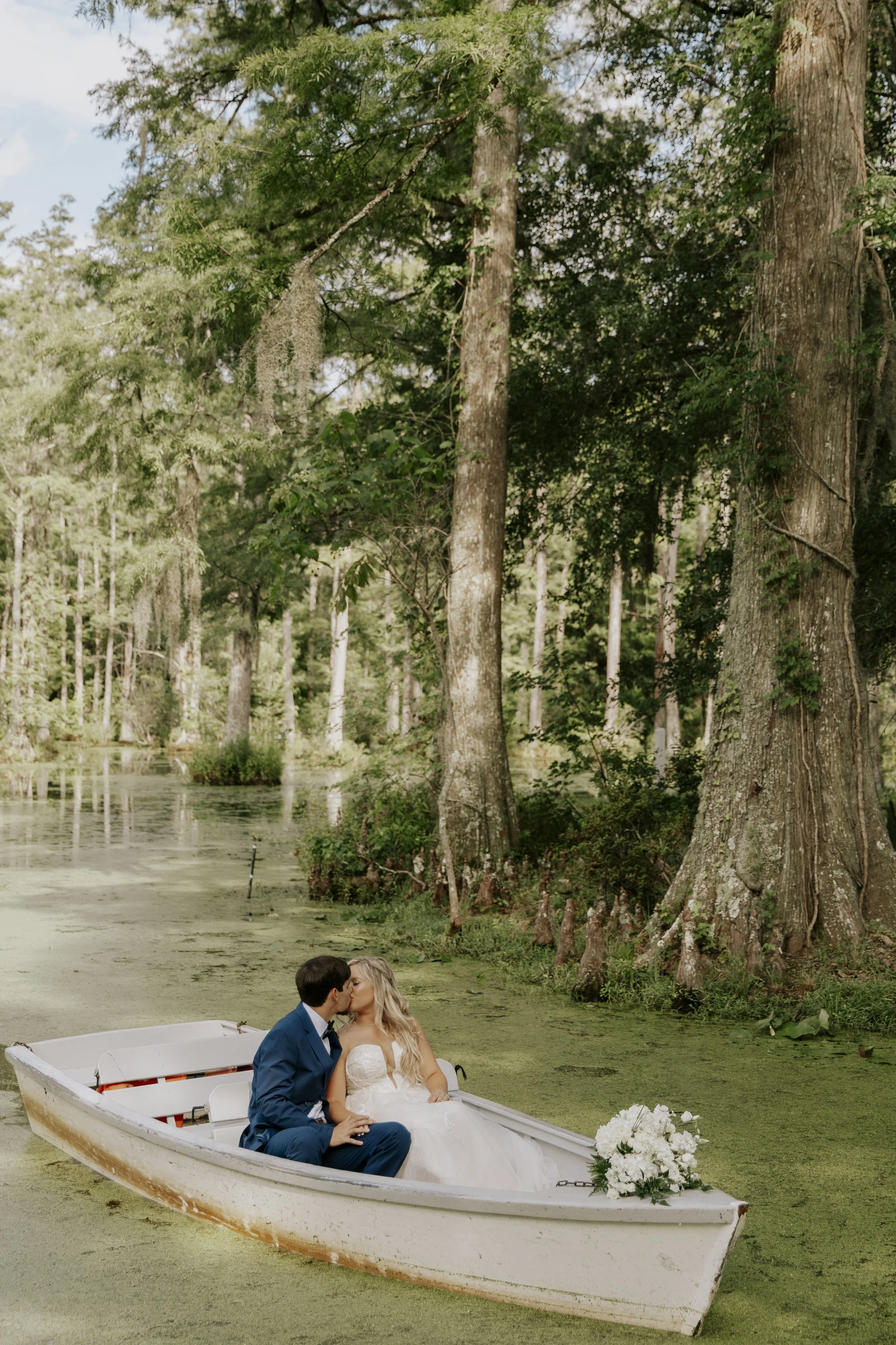 A bride and groom sitting in a small white boat with flowers on the front, kissing on a pond surrounded by tall trees and green foliage.