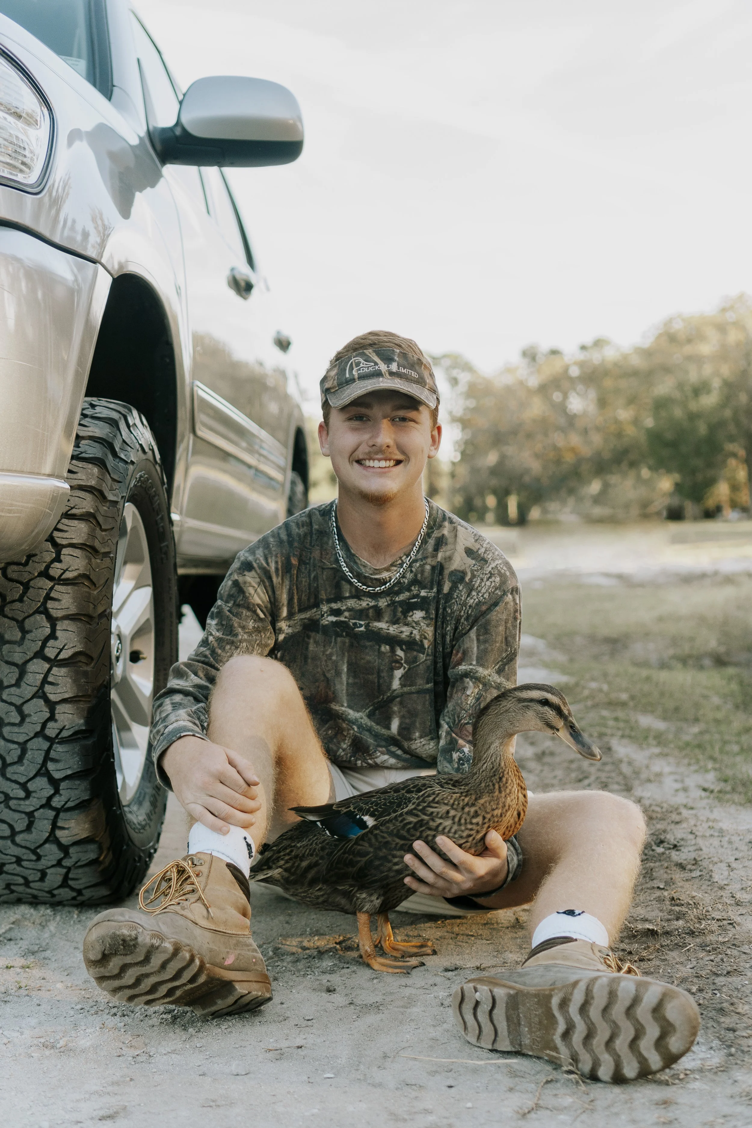 A smiling young man in camouflage shirt and tan boots sitting on the ground holding a duckling, with a duck in front of him, beside a silver truck, outdoors during daytime.