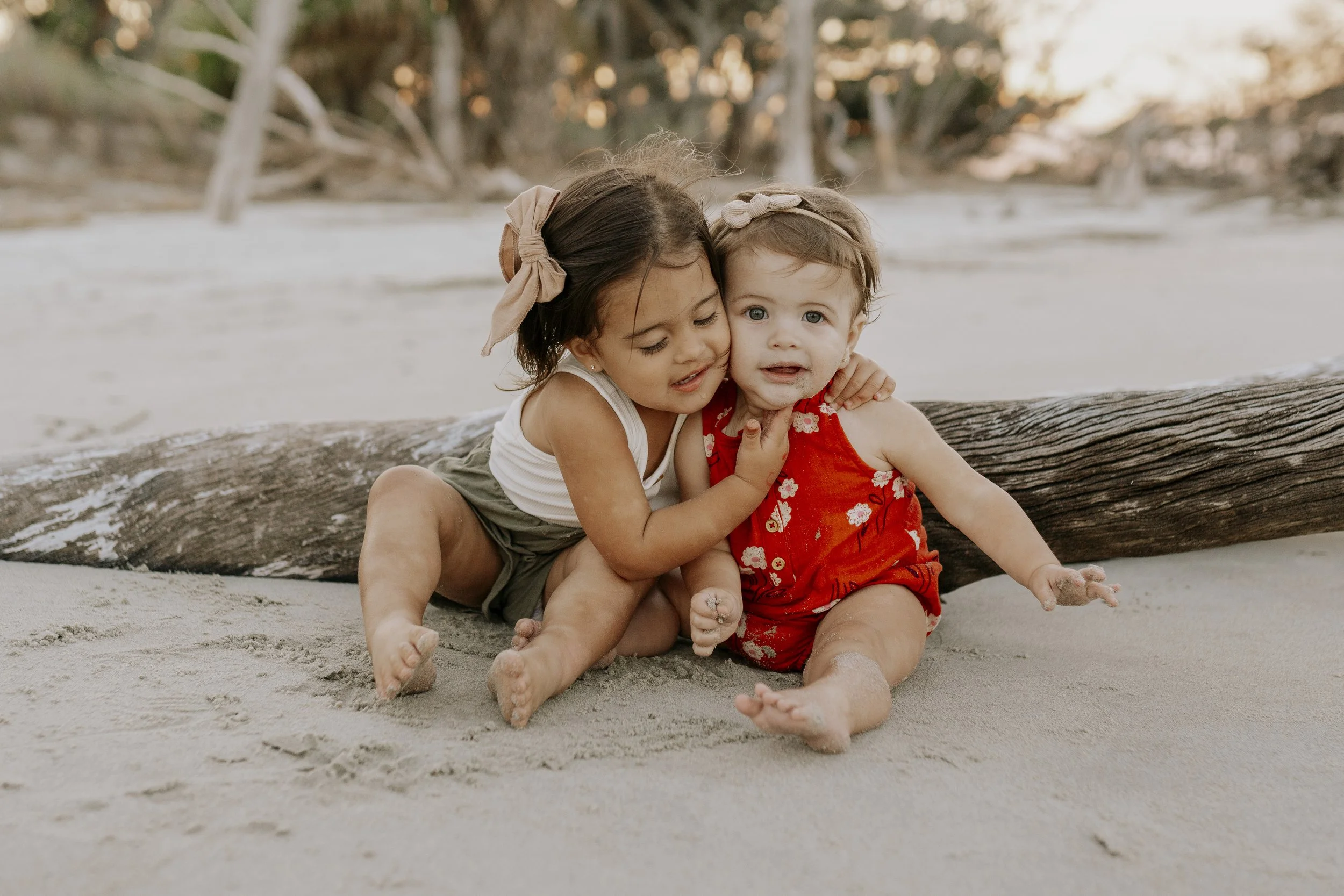 Two young girls sitting on the beach near a large fallen tree trunk, hugging and playing with sandy hands, during sunset with a background of trees.