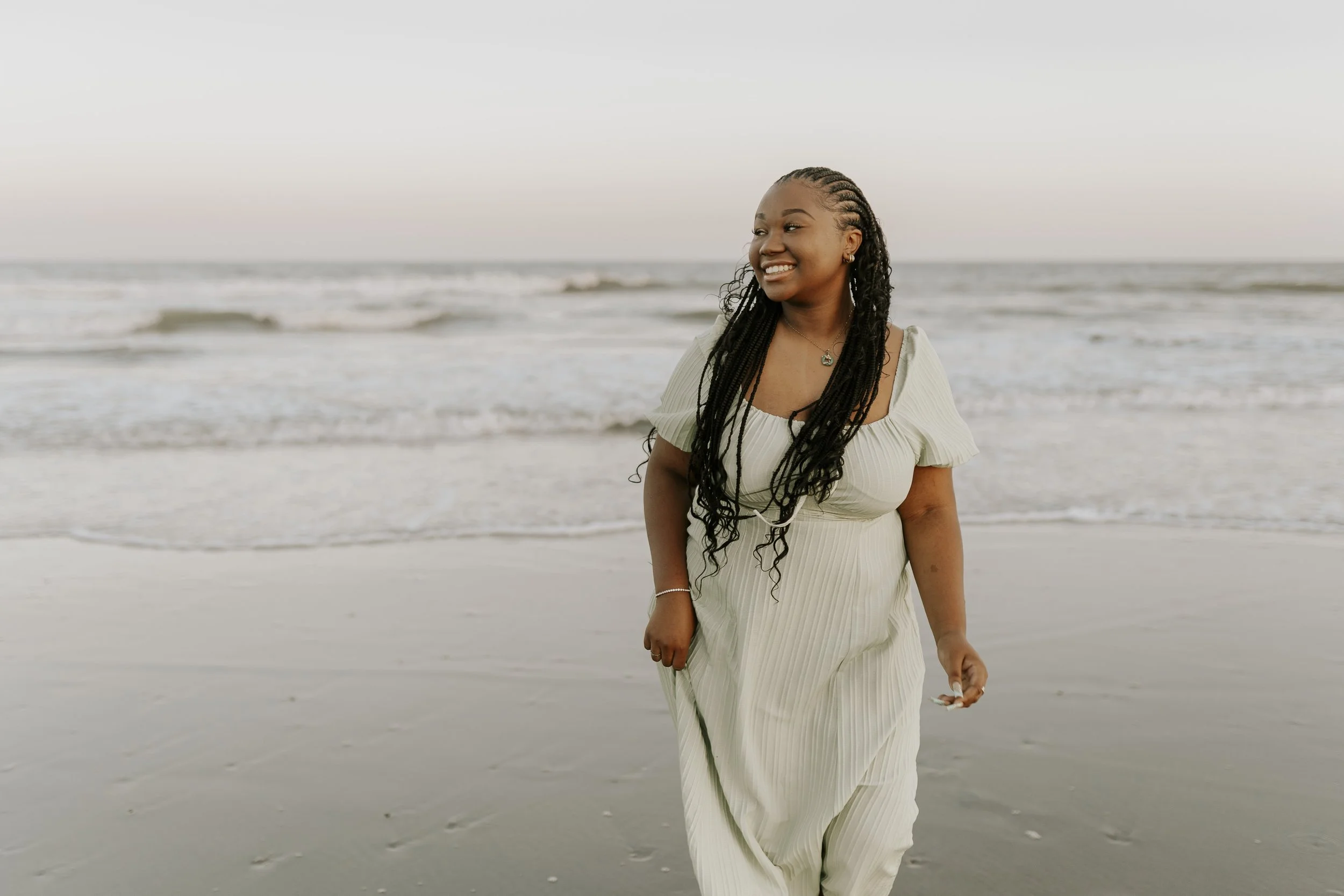 A woman with braided hair smiling at the beach, wearing a light-colored dress, with the ocean and a clear sky in the background.