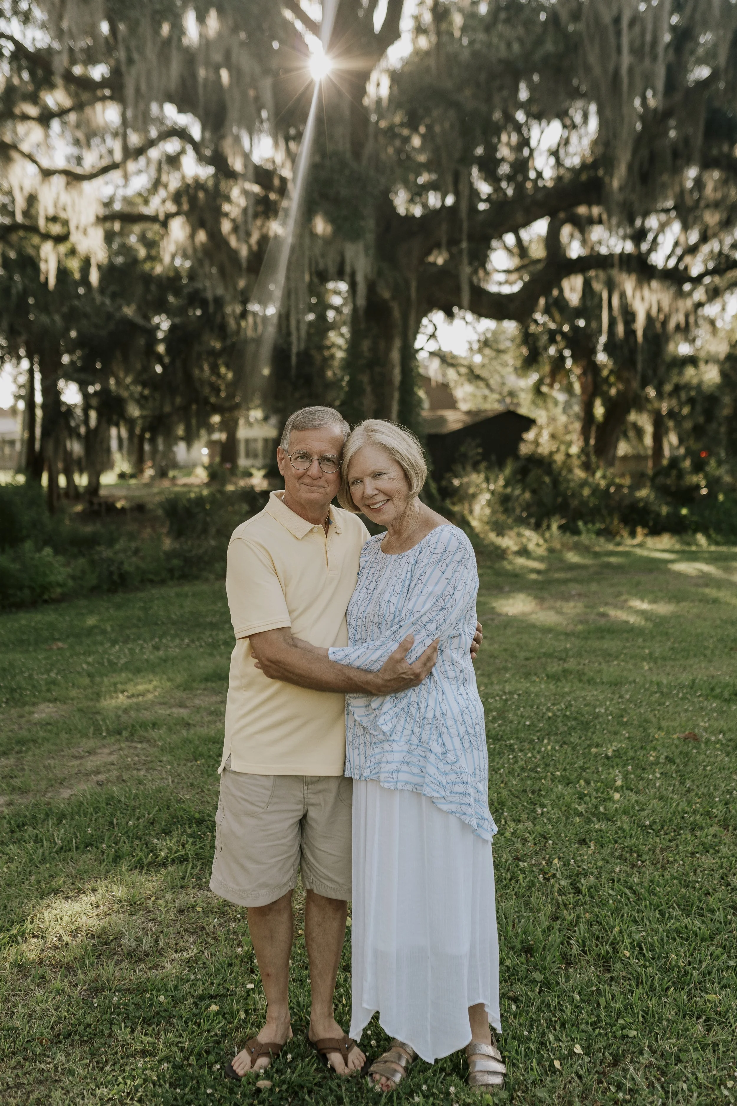 An elderly couple standing together outdoors in a park with large trees and Spanish moss. The man is wearing a yellow polo shirt and khaki shorts, while the woman is wearing a light blue patterned blouse and a long white skirt. They are embracing and smiling, with the sun shining through the trees in the background.