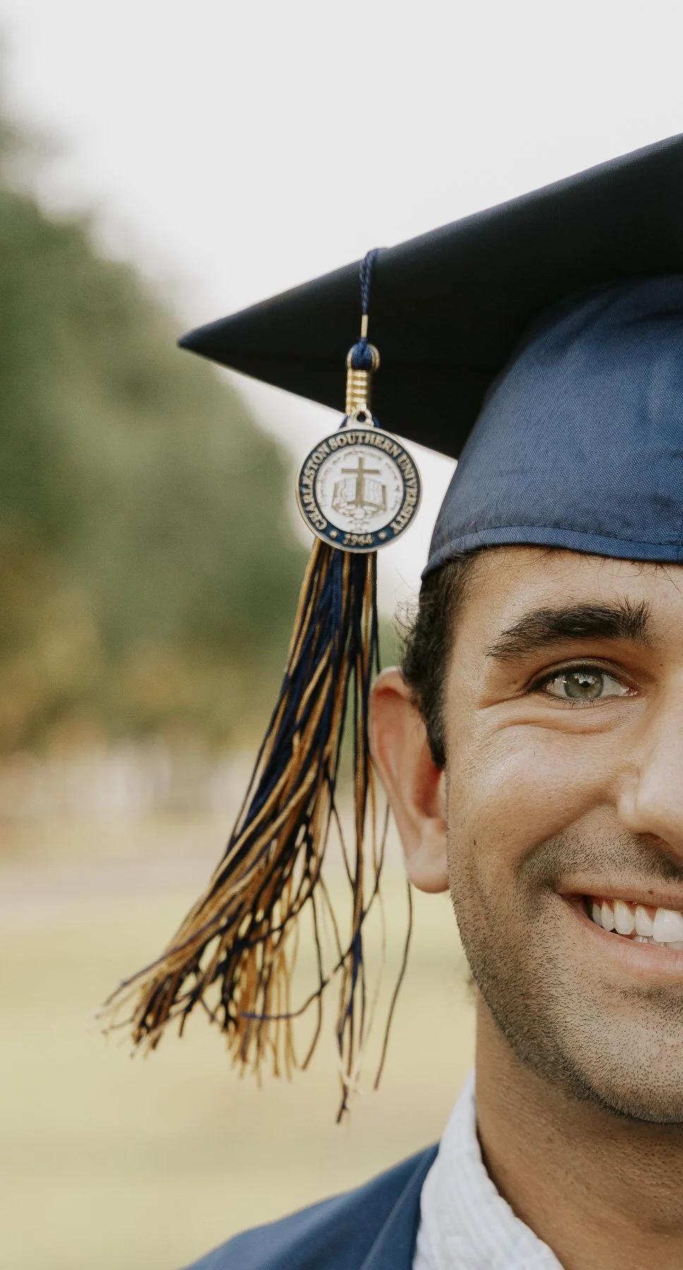Close-up of a young man wearing a graduation cap with a blue tassel and an emblem from a university, smiling outdoors.