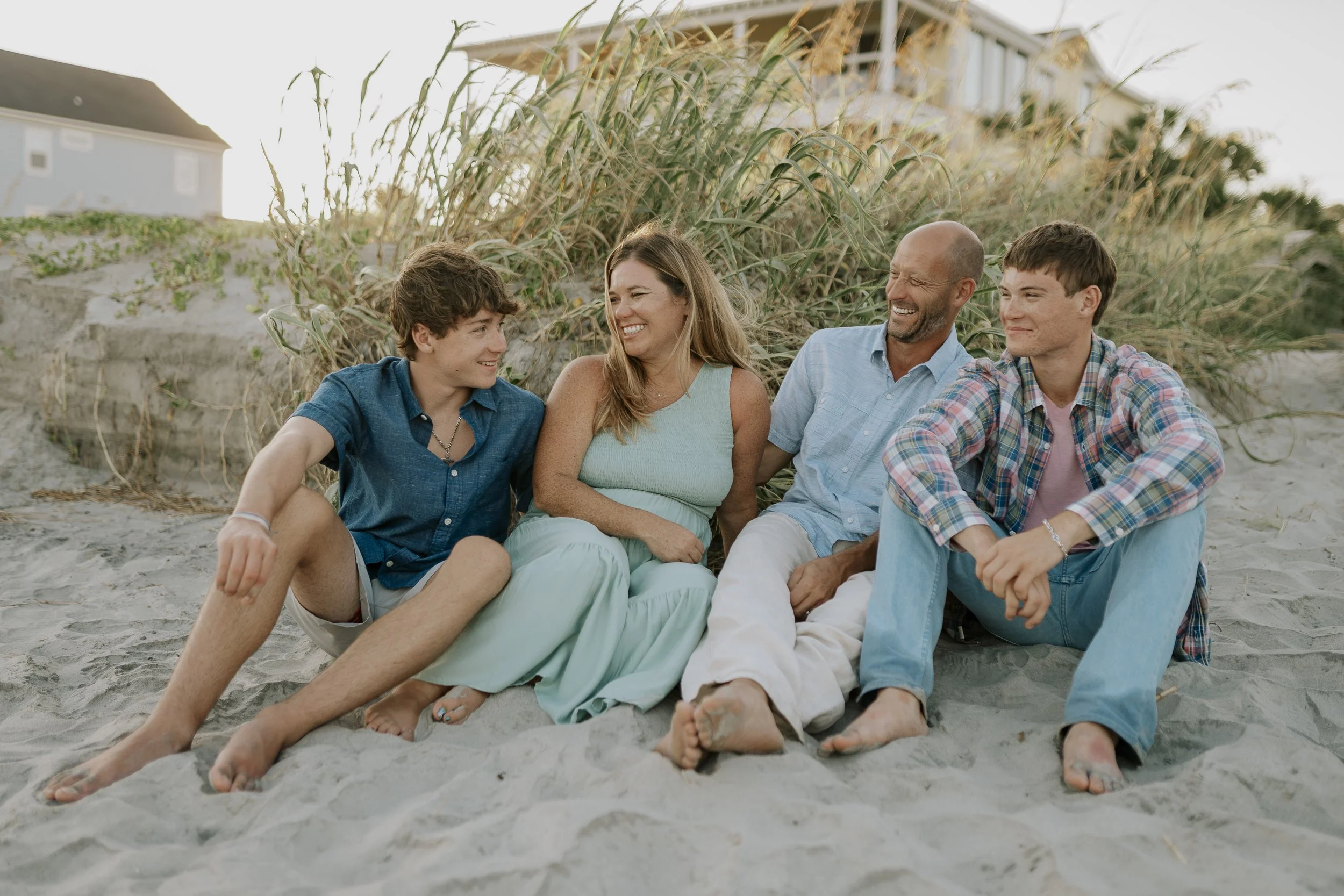 A family of four sitting together on the beach, smiling and enjoying the moment. The group includes two teenagers, a woman, and a man, all in casual summer clothing, with sand and beach grass in the background.