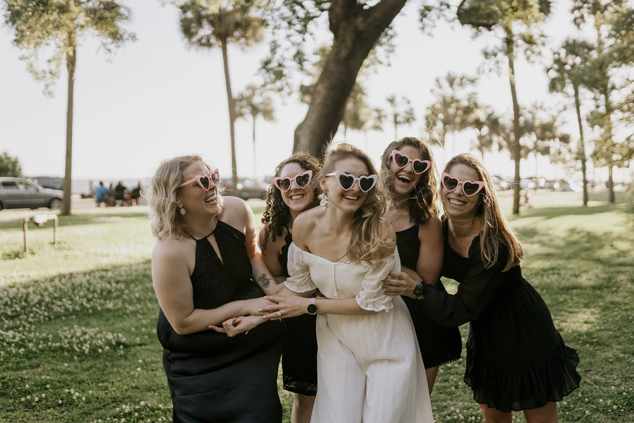 Group of five women outdoors, smiling and laughing, wearing heart-shaped sunglasses, with a large tree and parked cars in the background.