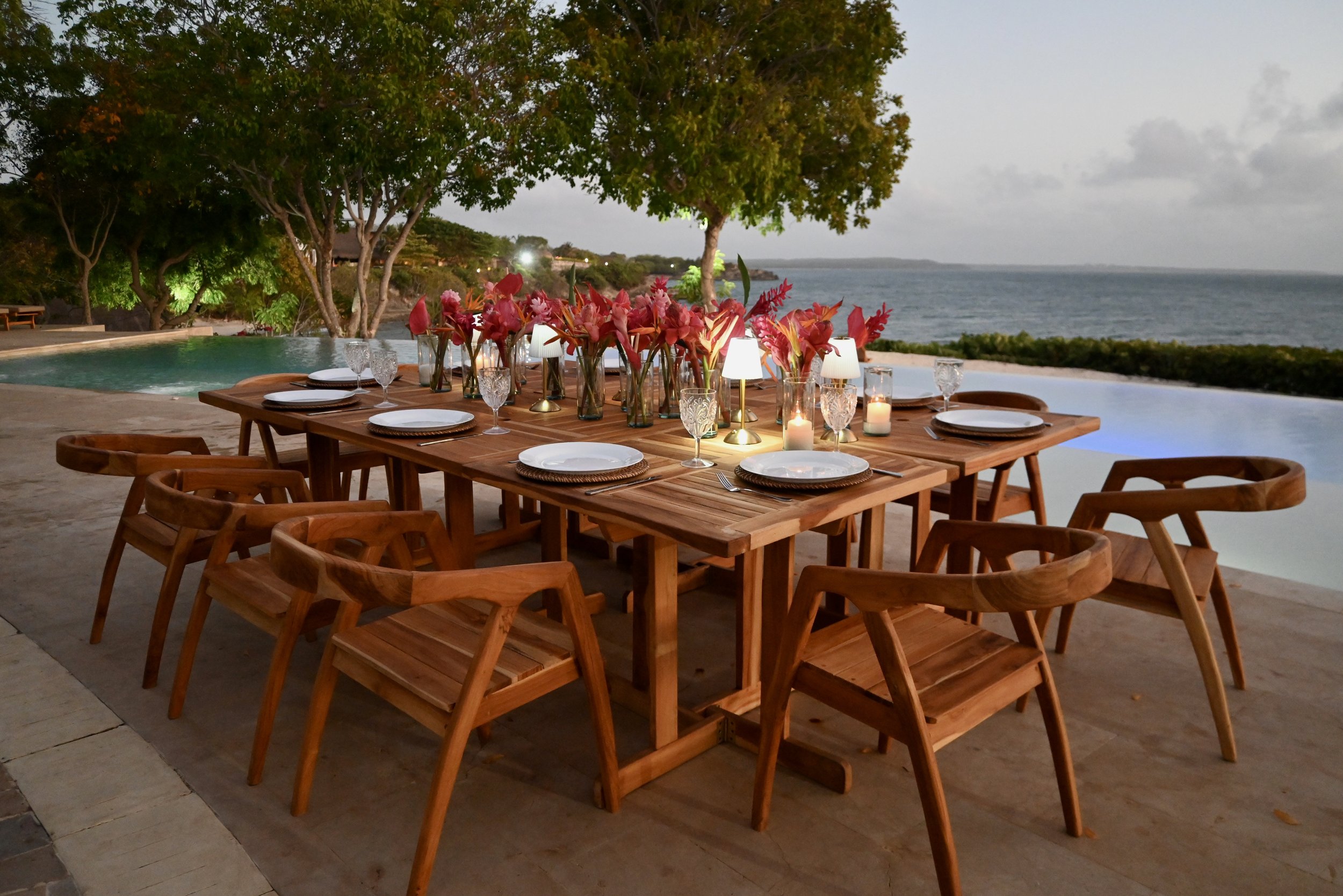 Mesa de decoración con flores y velas, vista al mar y árboles al atardecer.