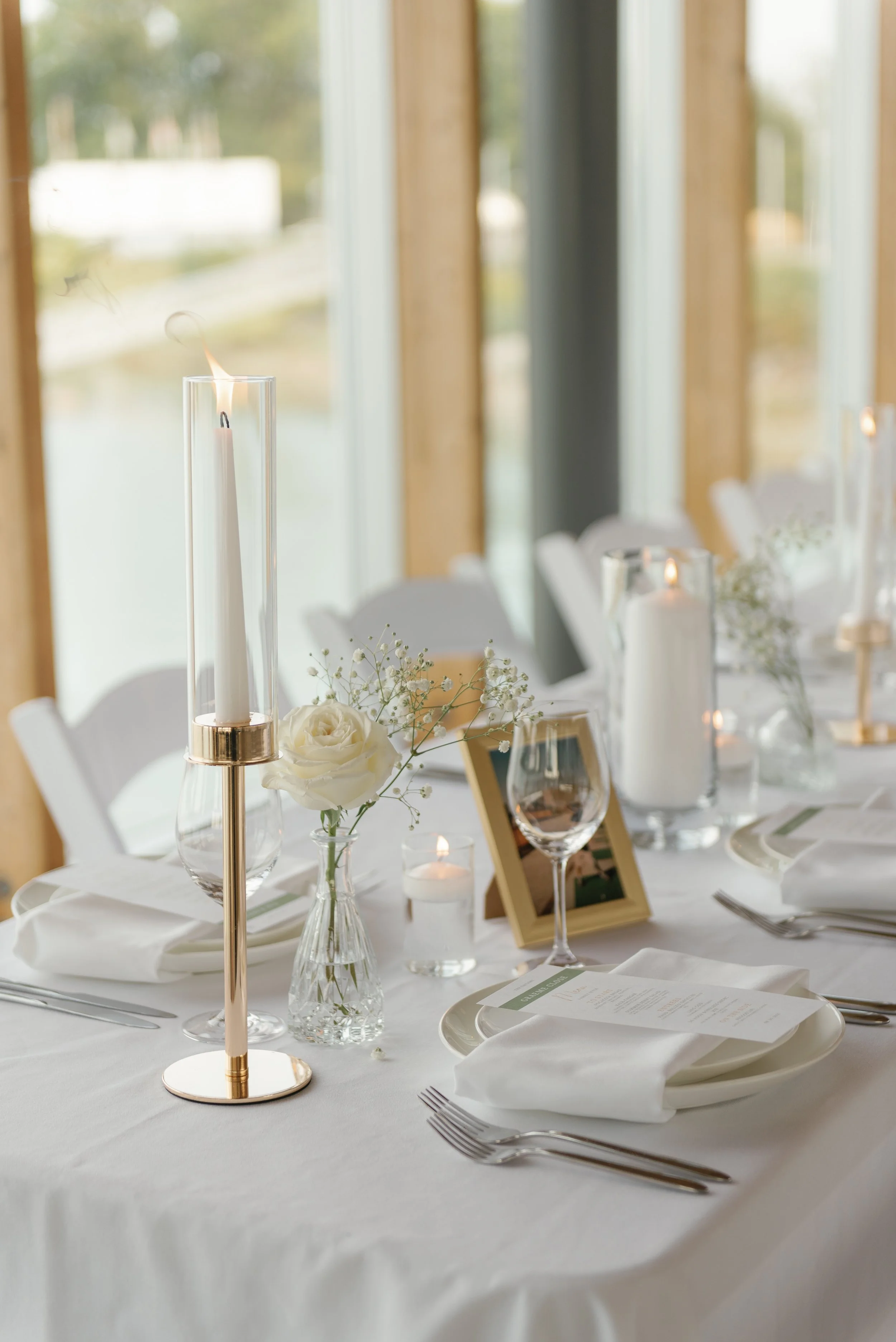 Elegantly set dining table with a tall candle holder, white roses, glassware, printed menu, and green framed photo, in a room with large windows and natural light.