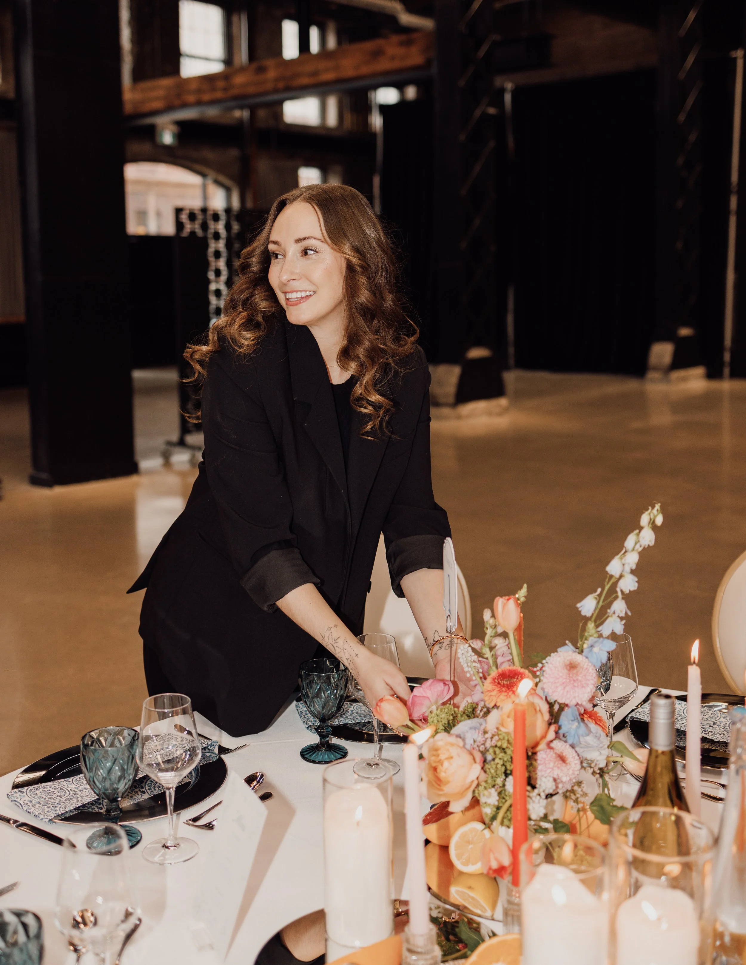 A woman with long, curly hair wearing a dark blazer is setting a dining table with floral arrangements and candles in a well-lit, rustic wooden interior.