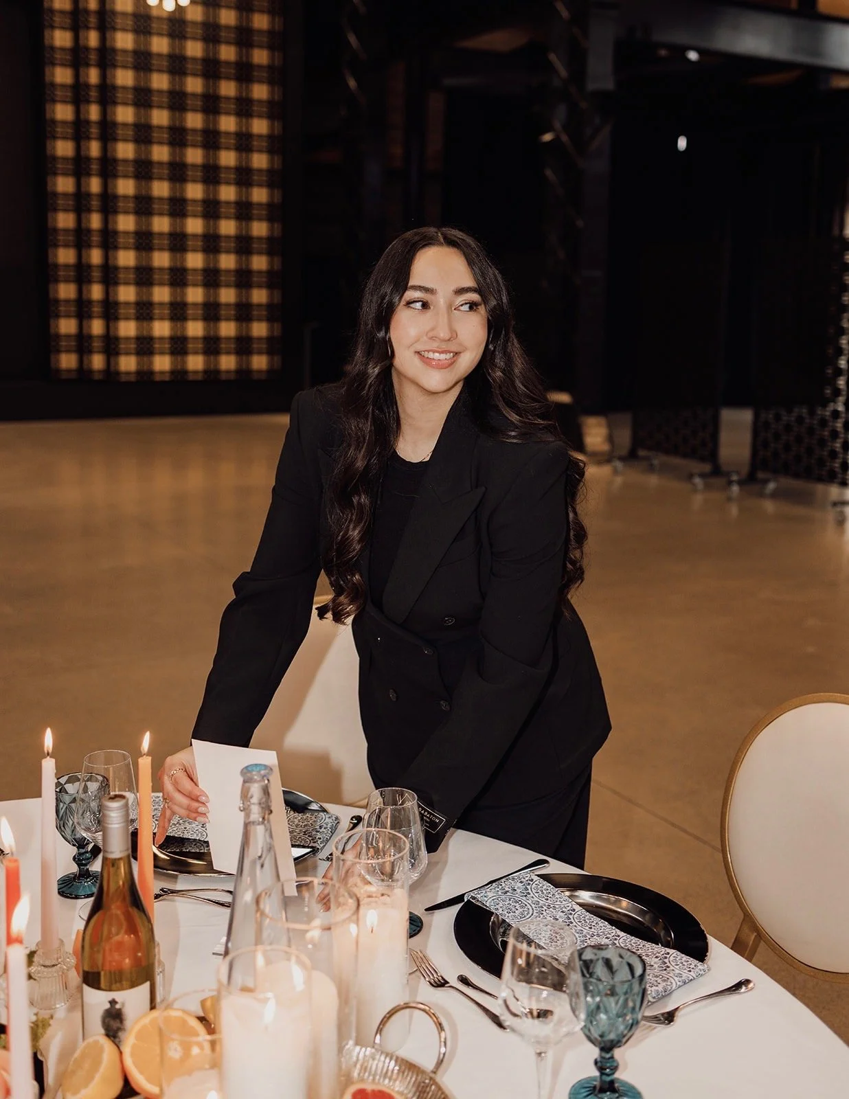 A woman in a black blazer leaning over a table set for a meal, with candles, wine glasses, and orange slices, in a dimly lit event space.