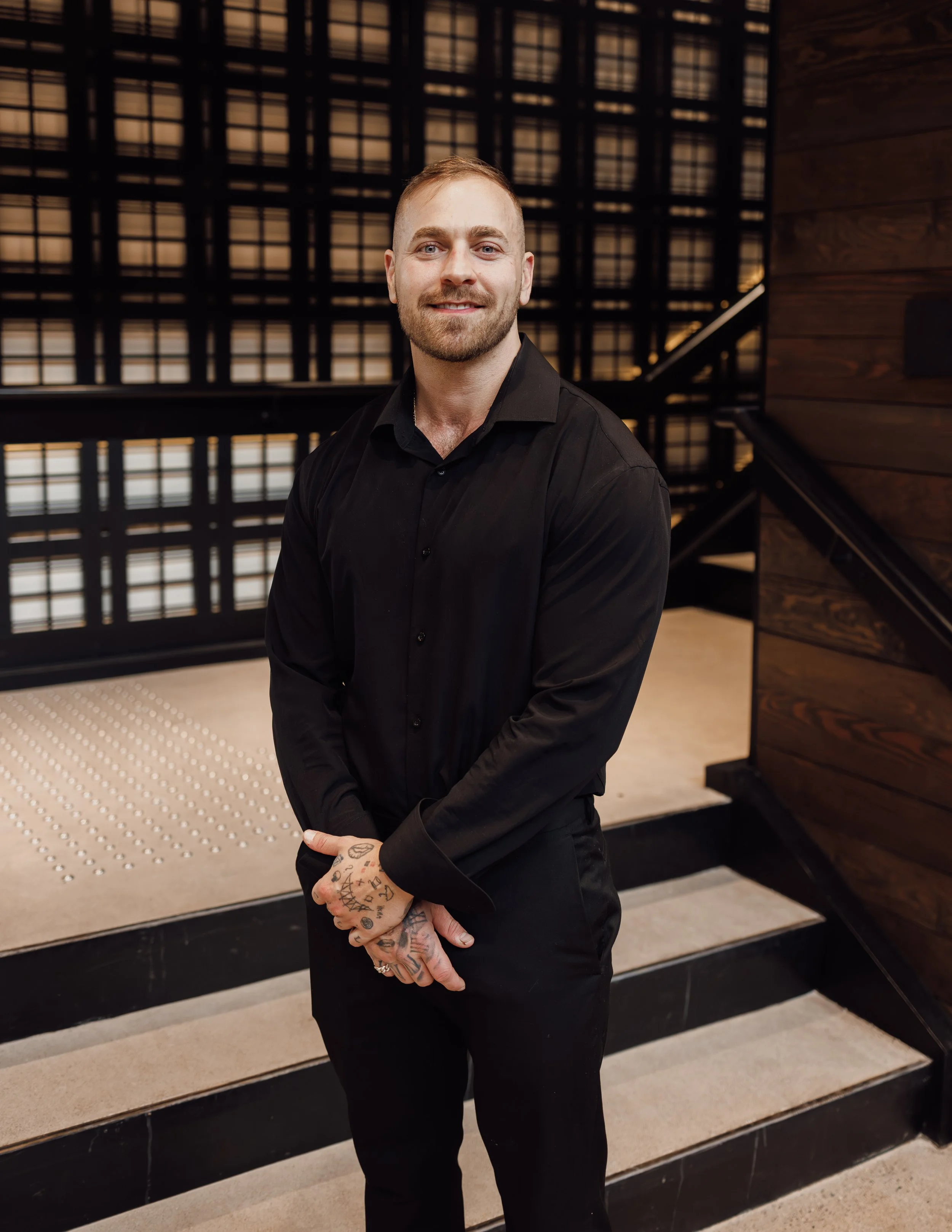 A smiling man with short hair, a beard, and tattoos on his hands wearing a black shirt and black pants, standing on a staircase with a modern wooden and metal design in the background.