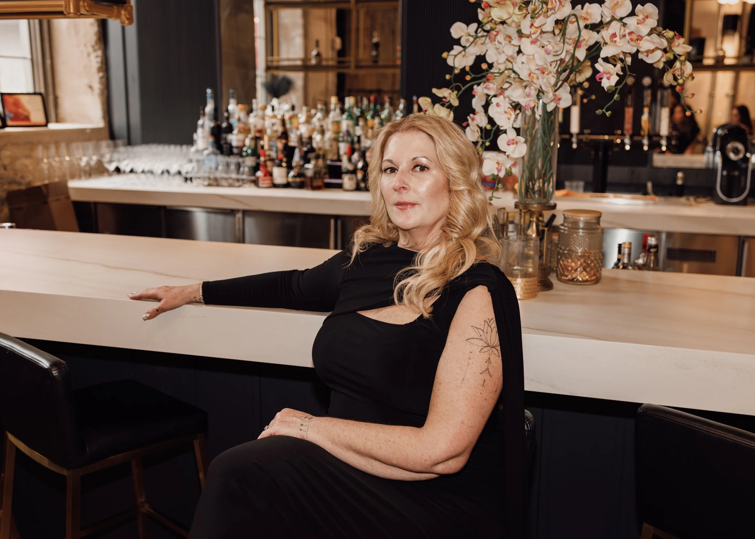 A woman with long blonde hair and tattoos, dressed in black, sitting at a bar with a necklace and jewelry, in a restaurant or bar with a large floral arrangement, bottles, and glasses in the background.