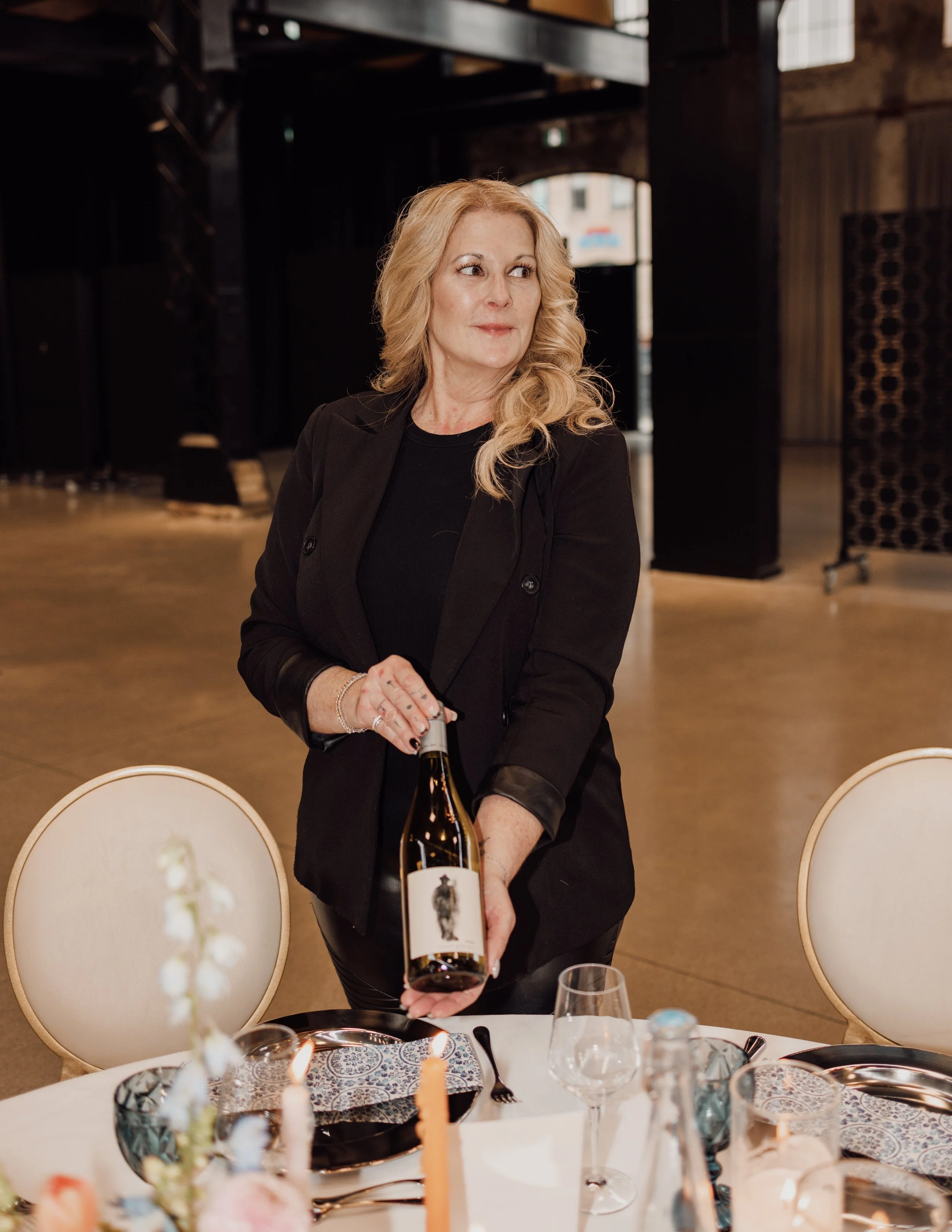 A woman with blonde hair dressed in black, standing at a dining table, holding a wine bottle, in an indoor setting with a dark background.
