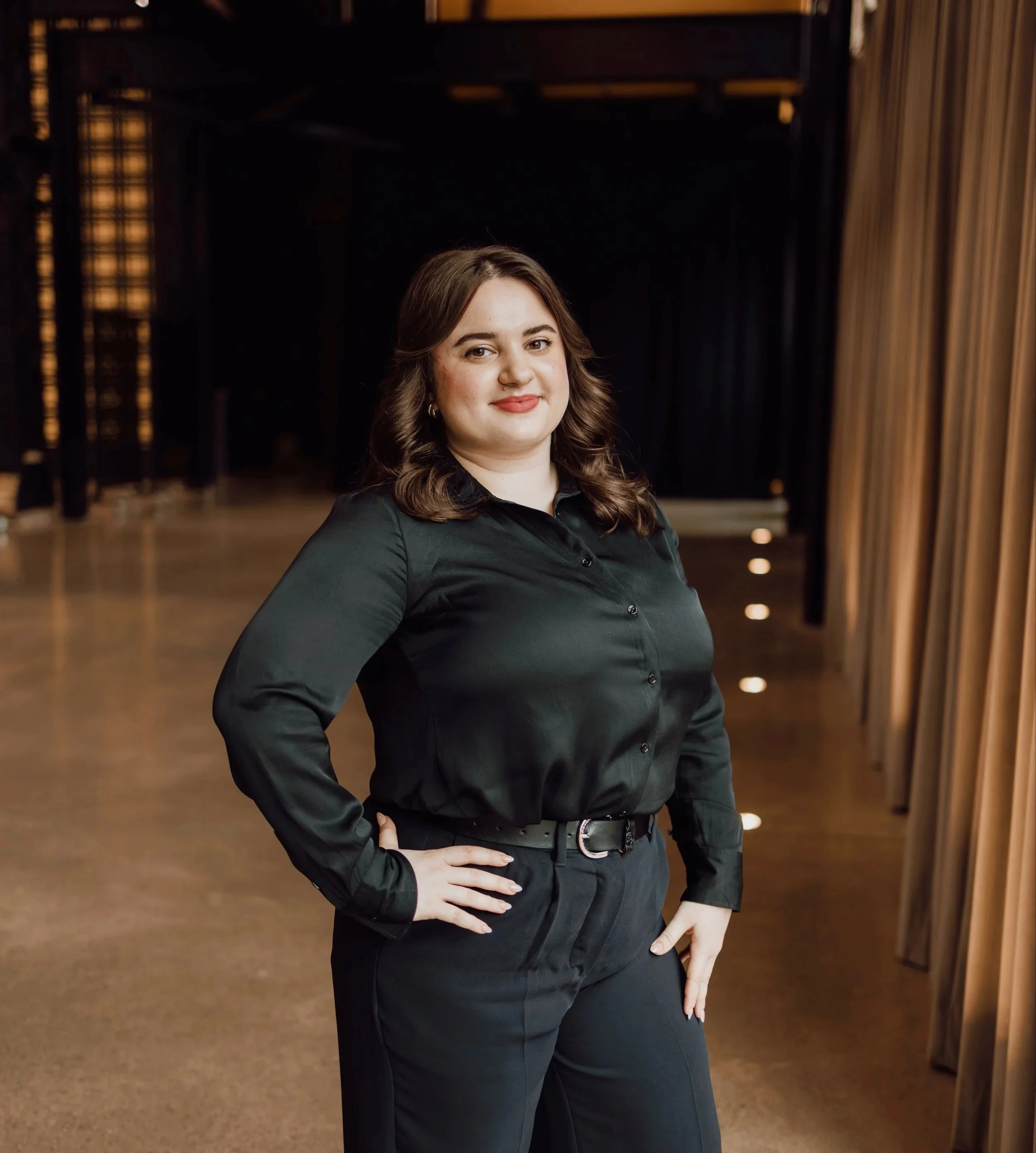 A woman with brown hair and makeup standing confidently indoors, wearing a black button-up shirt and black pants, with a brown curtain on one side and dark-colored furniture in the background.