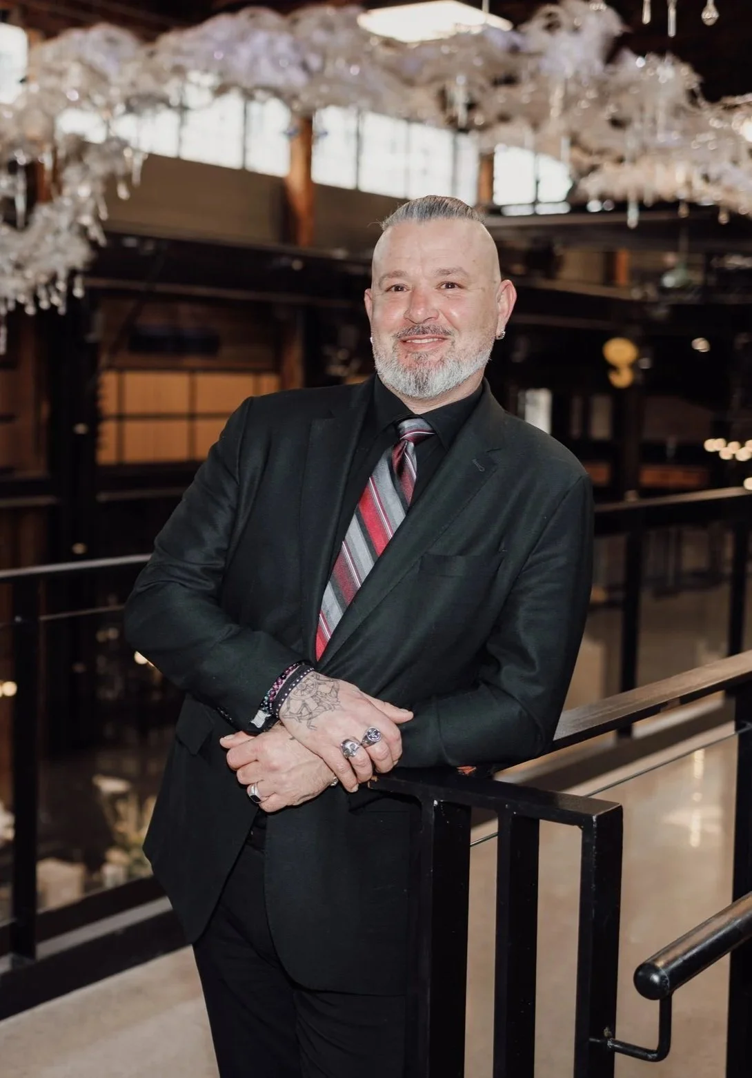 A man in a black suit with a red and gray striped tie, leaning on a railing inside a building with decorative lighting and wood accents, smiling at the camera.